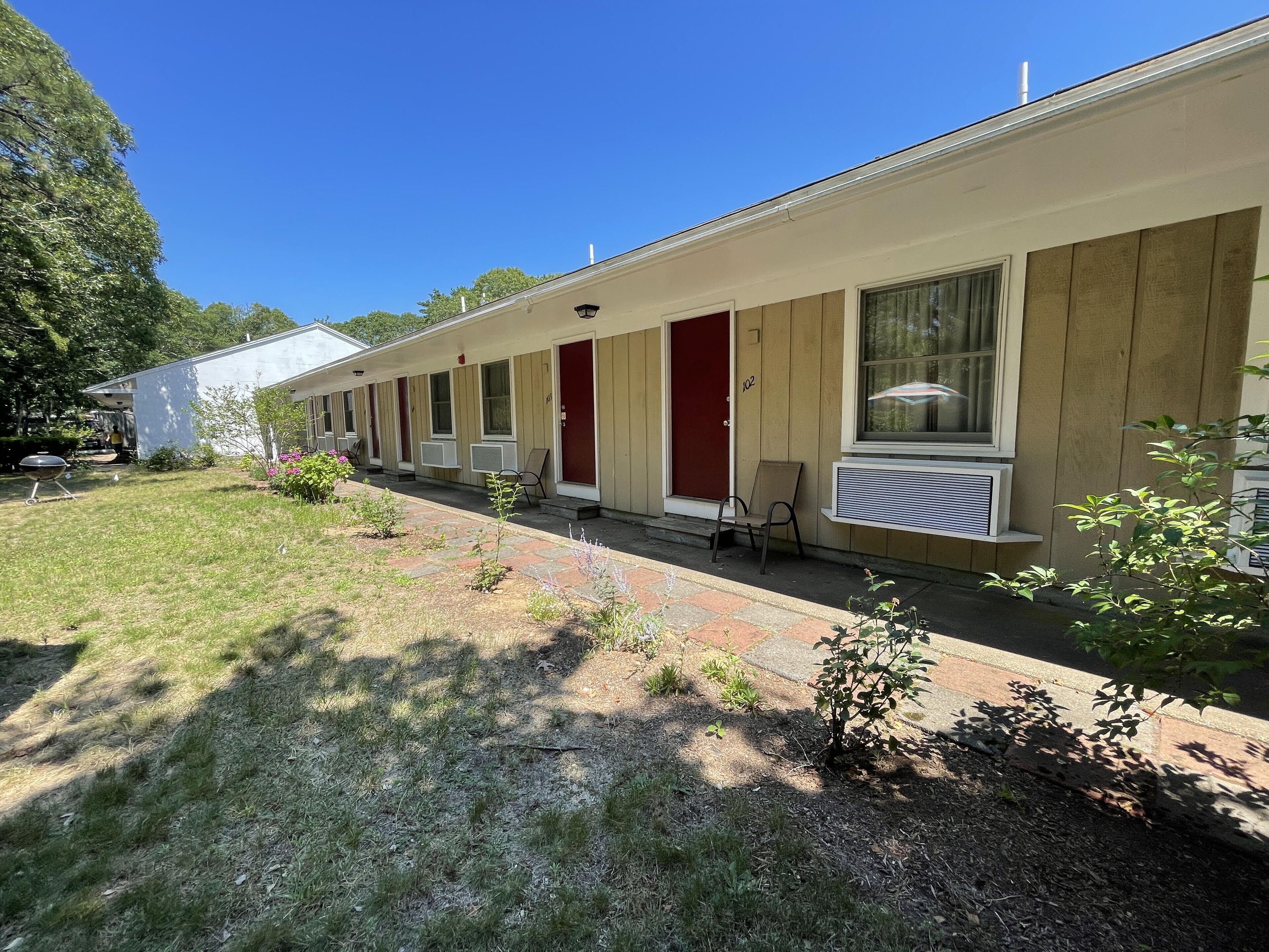 A row of single-story motel rooms with individual air conditioning units, a small garden area, and a clear blue sky.