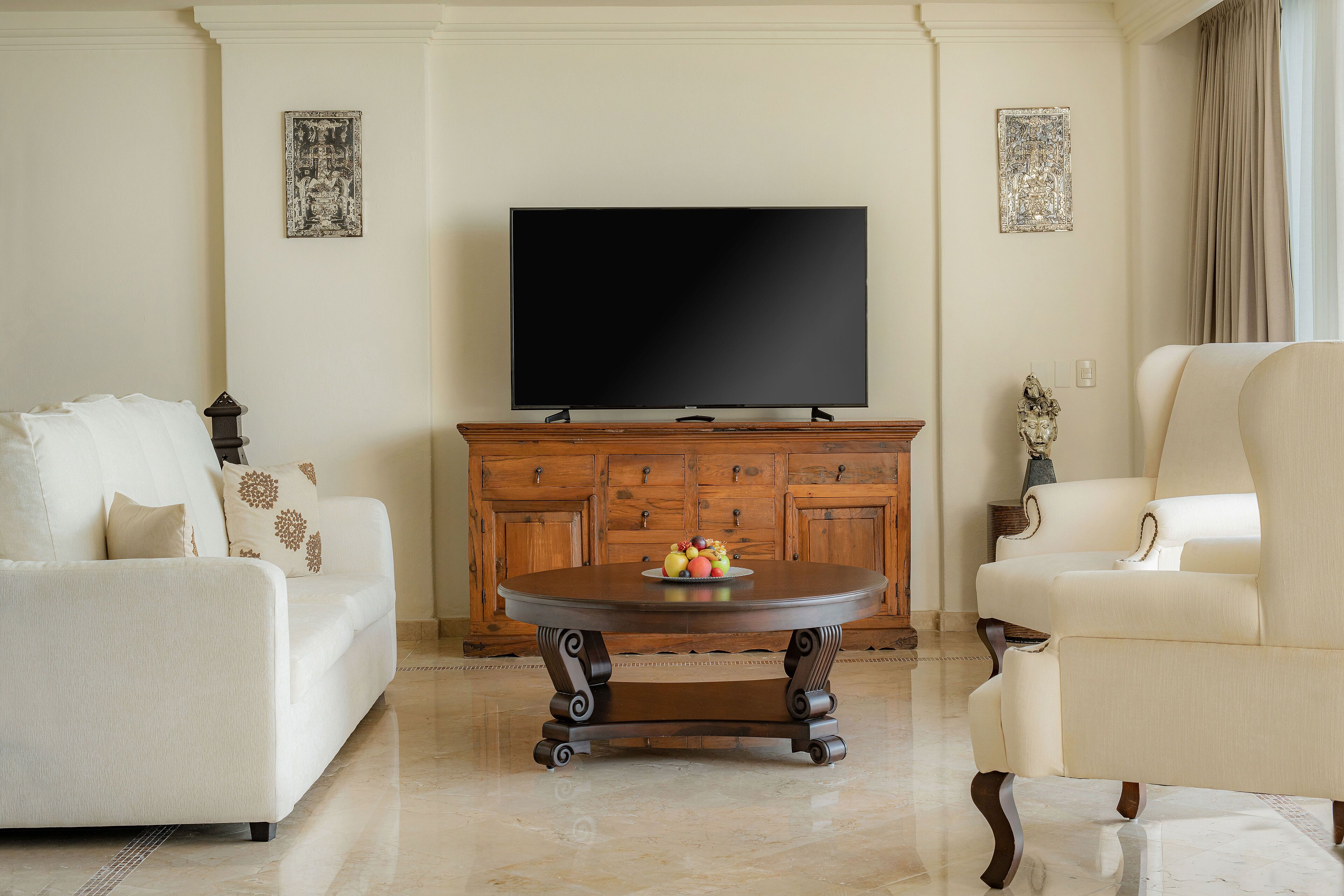Una sala de estar con una televisión de pantalla plana, una mesa de centro de madera con fruta, un sofá blanco y un sillón color crema.
