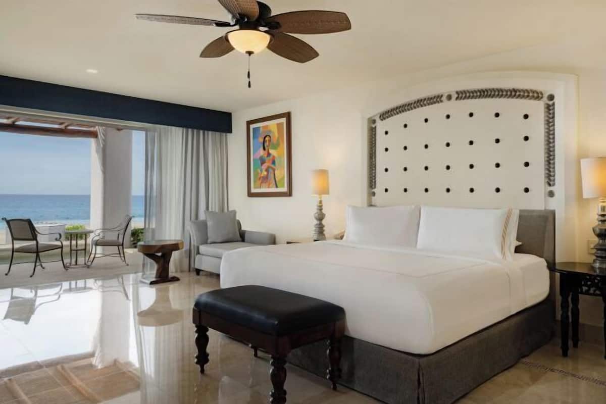 Ein Hotelzimmer mit einem großen Bett, einem Deckenventilator, Meerblick, einem Gemälde an der Wand und einem Essbereich mit Stühlen und Tisch.