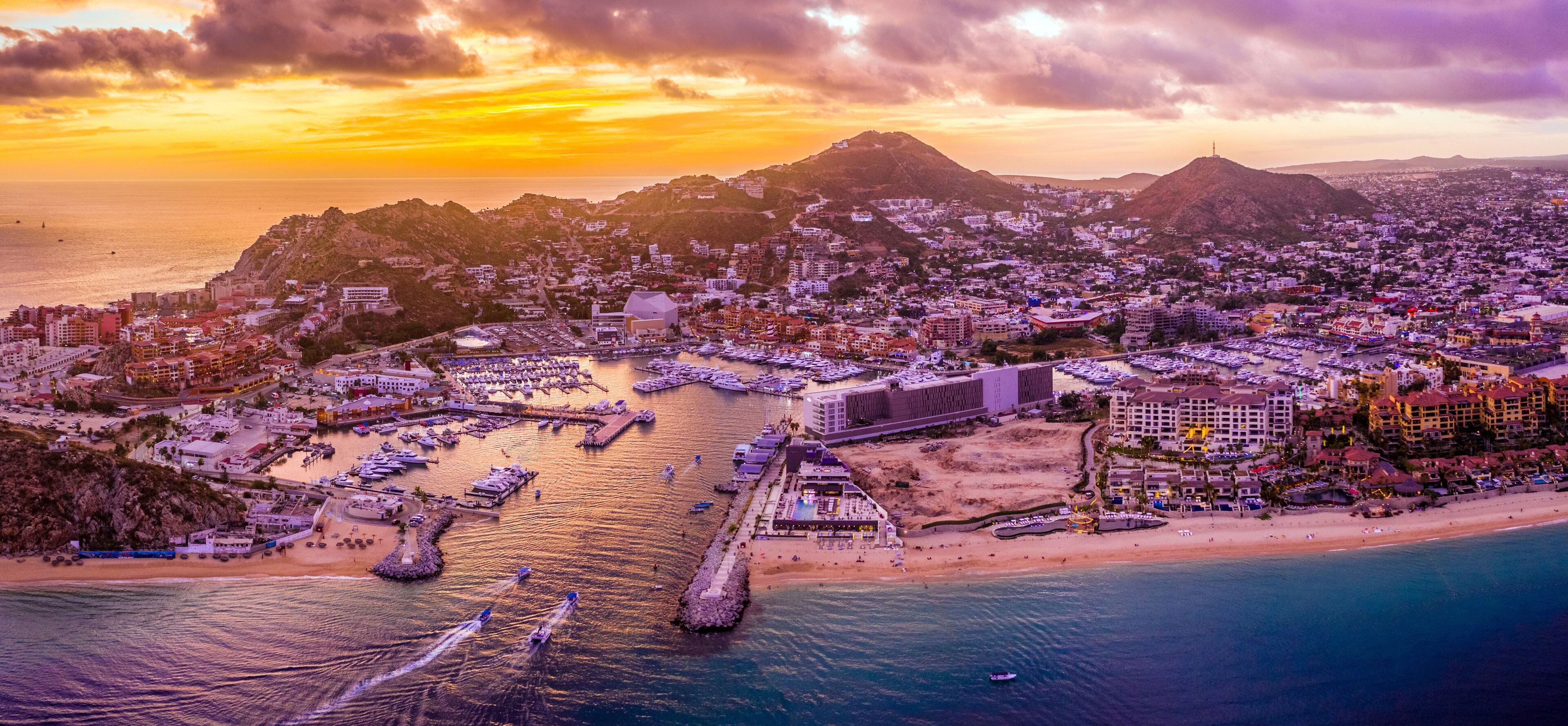 An aerial view of a coastal city with a marina, beach, and mountainous backdrop during sunset.