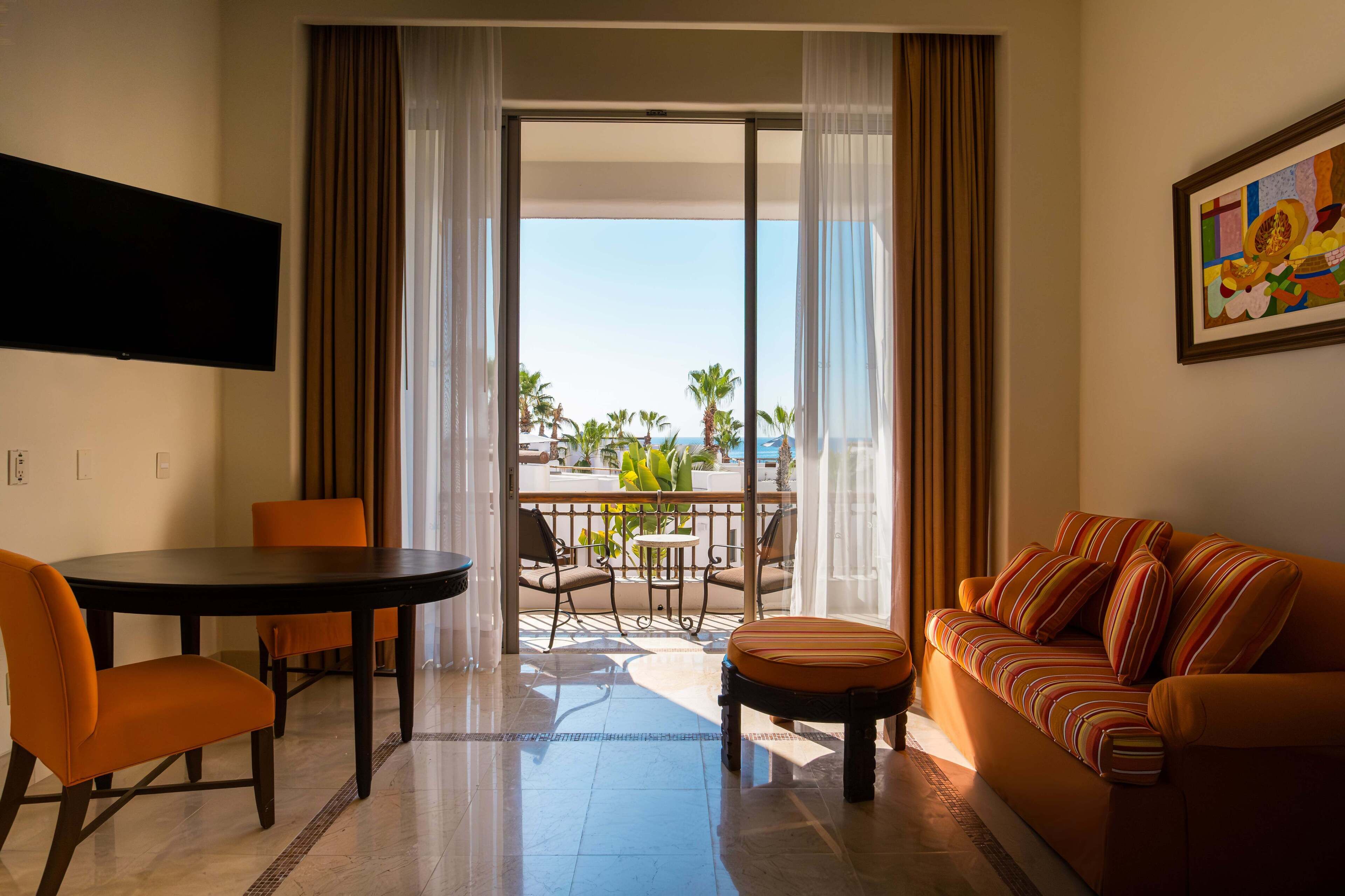 Habitación de hotel con balcón, una mesa de comedor con sillas, un sofá y un televisor de pantalla plana montado en la pared.