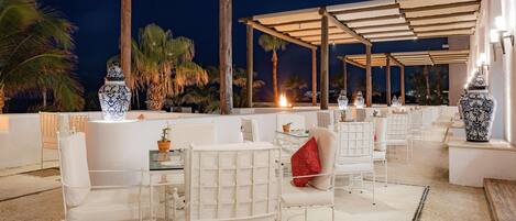 Un área de estar al aire libre con techo, con sillas y mesas blancas, rodeada de palmeras y jarrones decorativos.