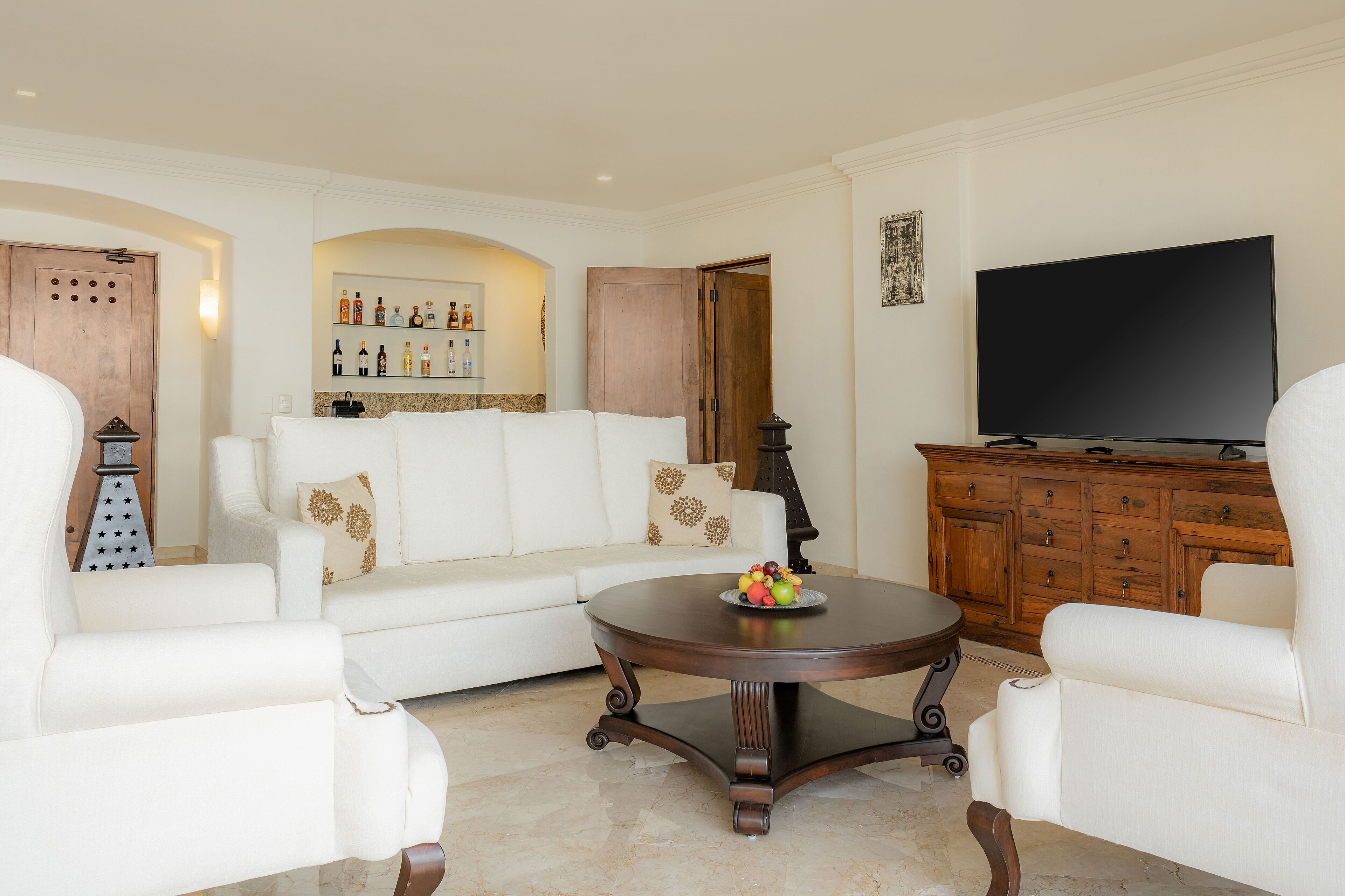 Uma sala de estar com sofá branco, mesa de centro redonda com frutas, televisão e um armário de madeira.
