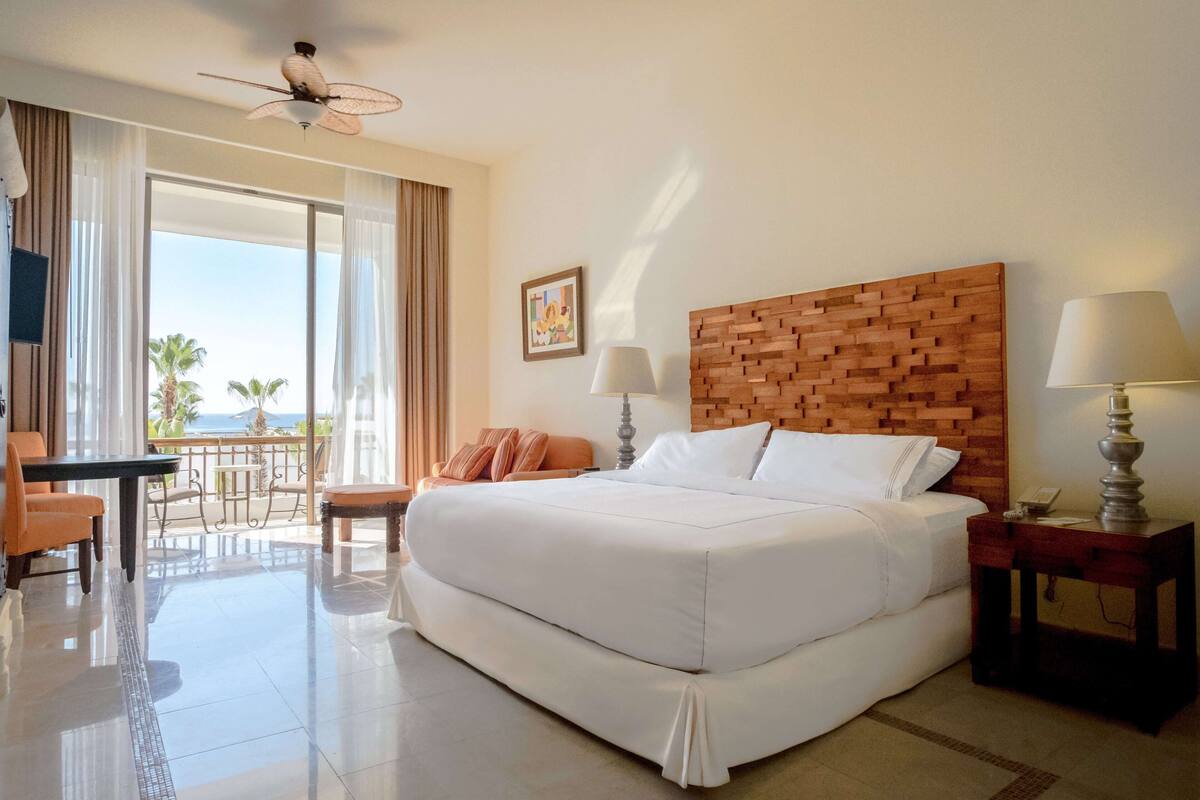 Ein Hotelzimmer mit einem großen Bett, einem Holz-Kopfteil, zwei Nachttischen mit Lampen, einem Schreibtisch mit Stuhl sowie einem Balkon mit Blick auf Palmen und das Meer.