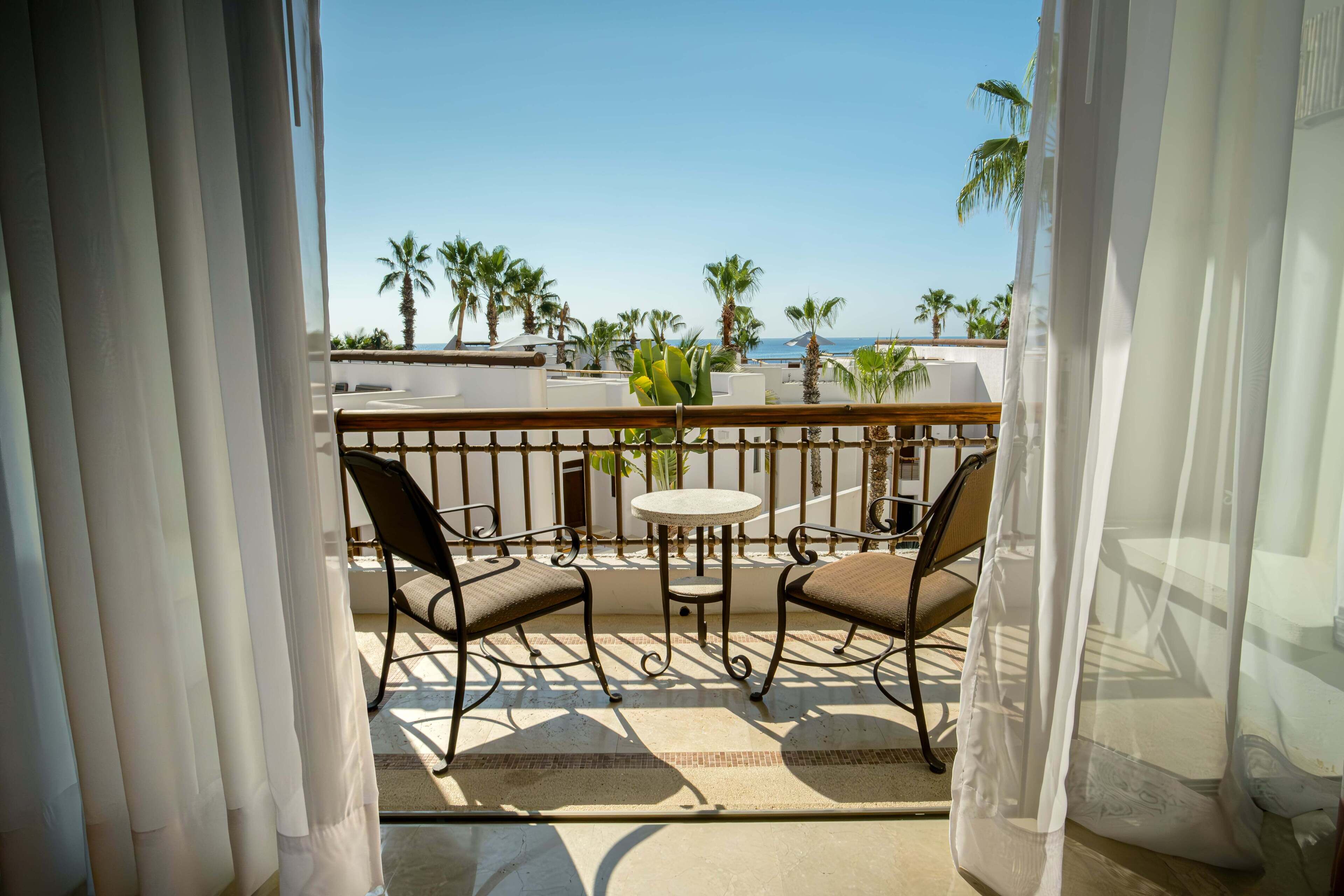 A balcony with two chairs and a table, overlooking palm trees and a clear sky.