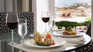 Une table dressée avec des assiettes de nourriture, des verres de vin, et une vue sur une plage et des chaises.