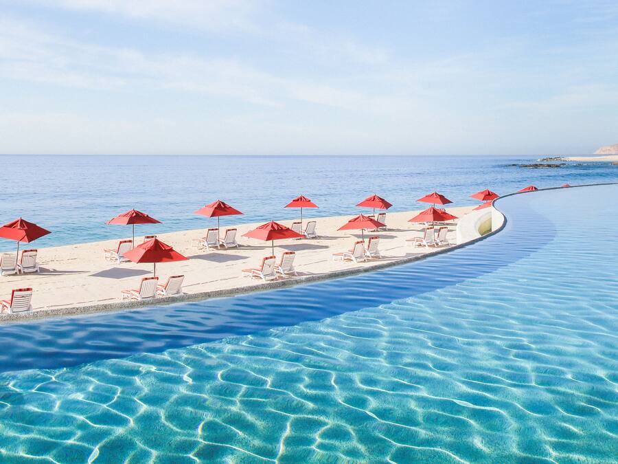 A poolside area with red umbrellas and white lounge chairs, overlooking a beach.