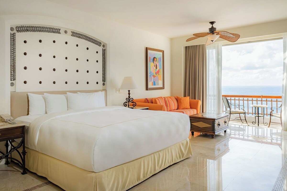Une chambre d’hôtel avec un grand lit, un canapé, un balcon donnant sur la mer, un ventilateur de plafond et un tableau au mur.