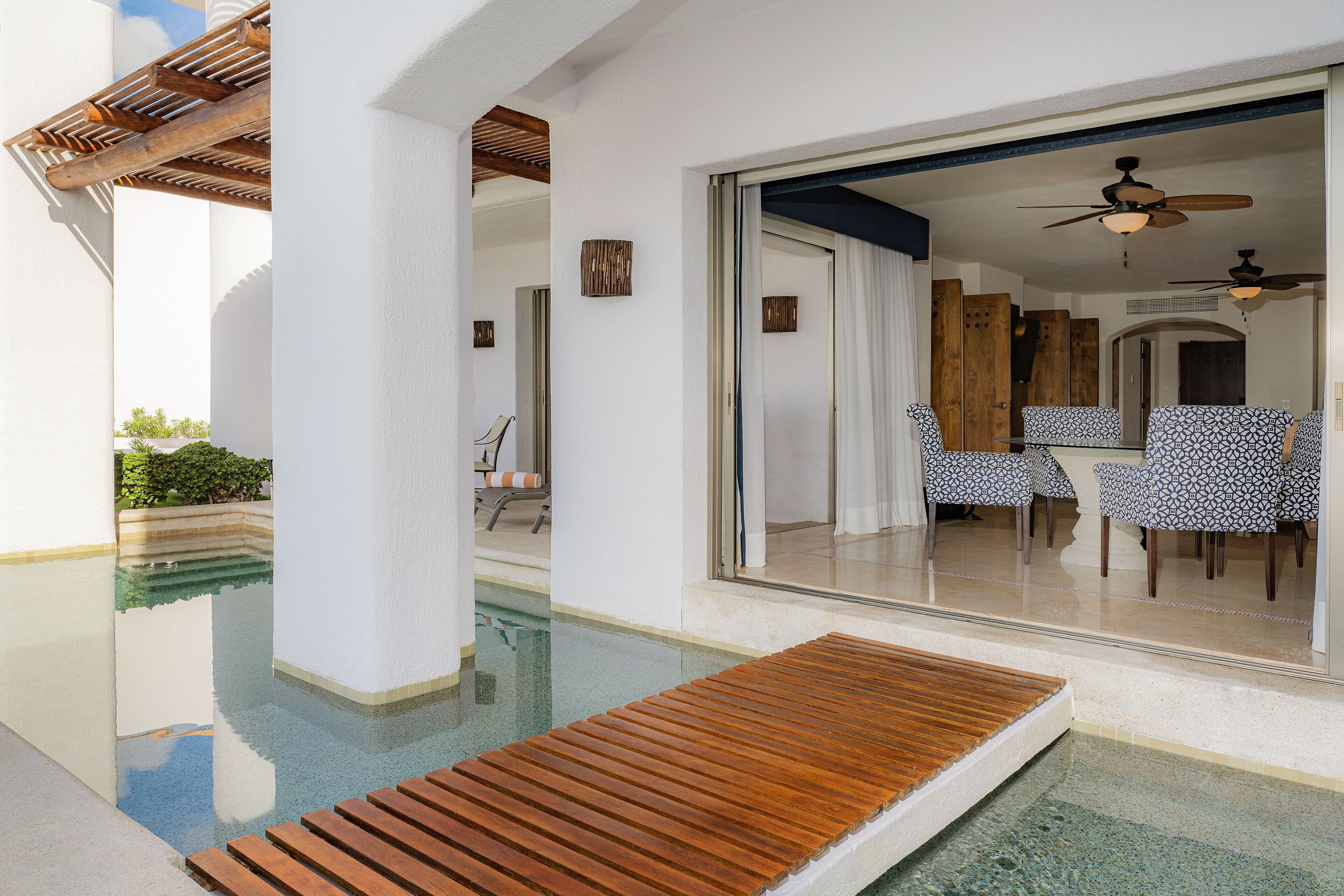 Un área de piscina con terraza de madera, zona de comedor con sillas estampadas y un ventilador de techo.