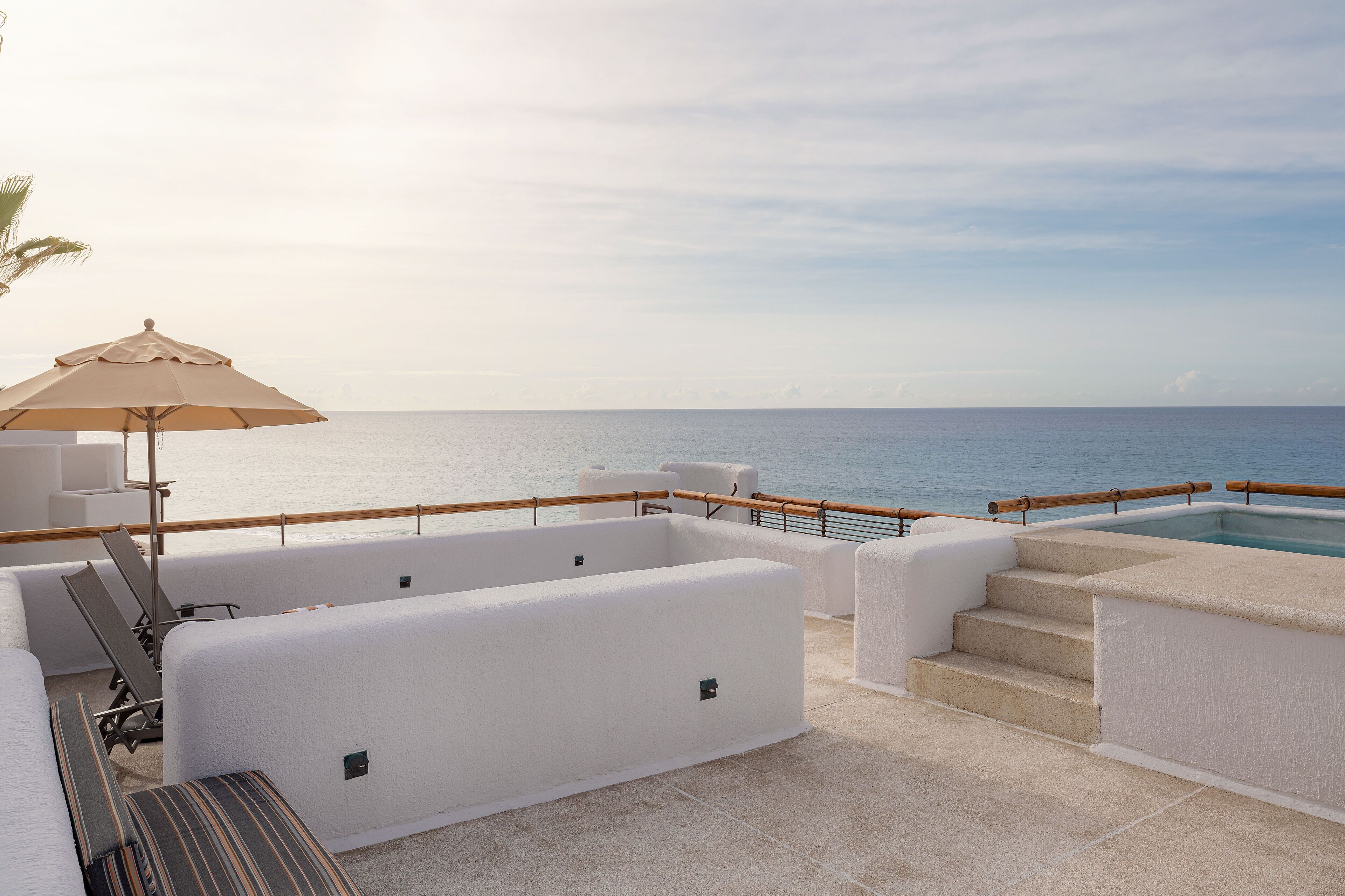 Zona de estar al aire libre iluminada por el sol, con piscina, sillones reclinables blancos con cojines y barandilla de madera con vista al mar.