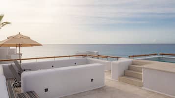 Zona de estar al aire libre iluminada por el sol, con piscina, sillones reclinables blancos con cojines y barandilla de madera con vista al mar.