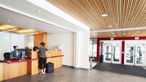 A reception desk with a computer, a counter, and a person standing beside it. There is a suitcase on the floor and a door with an exit sign. - University of Calgary - Seasonal Residence (Calgary)