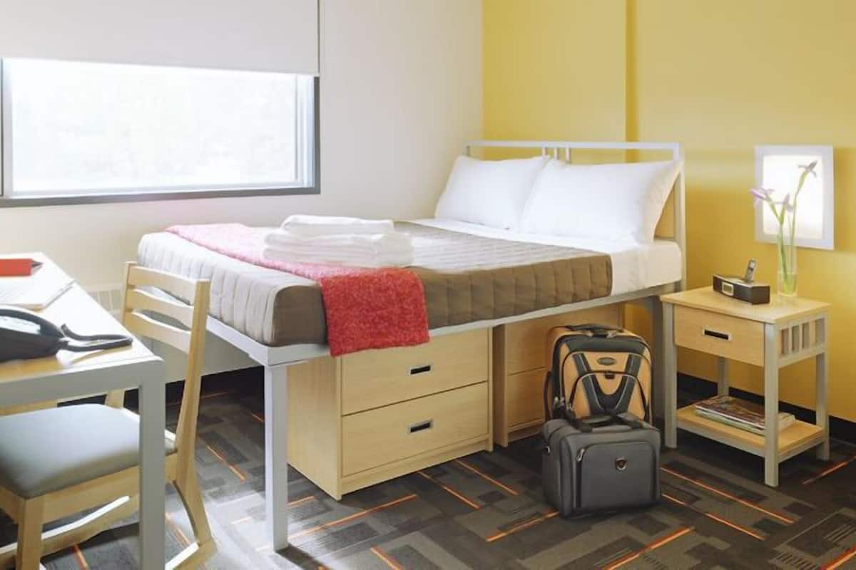 A hotel room with a single bed, a desk, a chair, a small table, a lamp, and a suitcase.