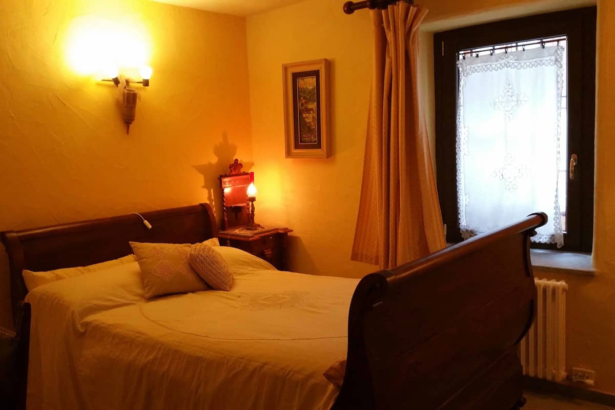 A bedroom with a wooden bed, a nightstand with a lamp, a framed picture on the wall, and a window with curtains.