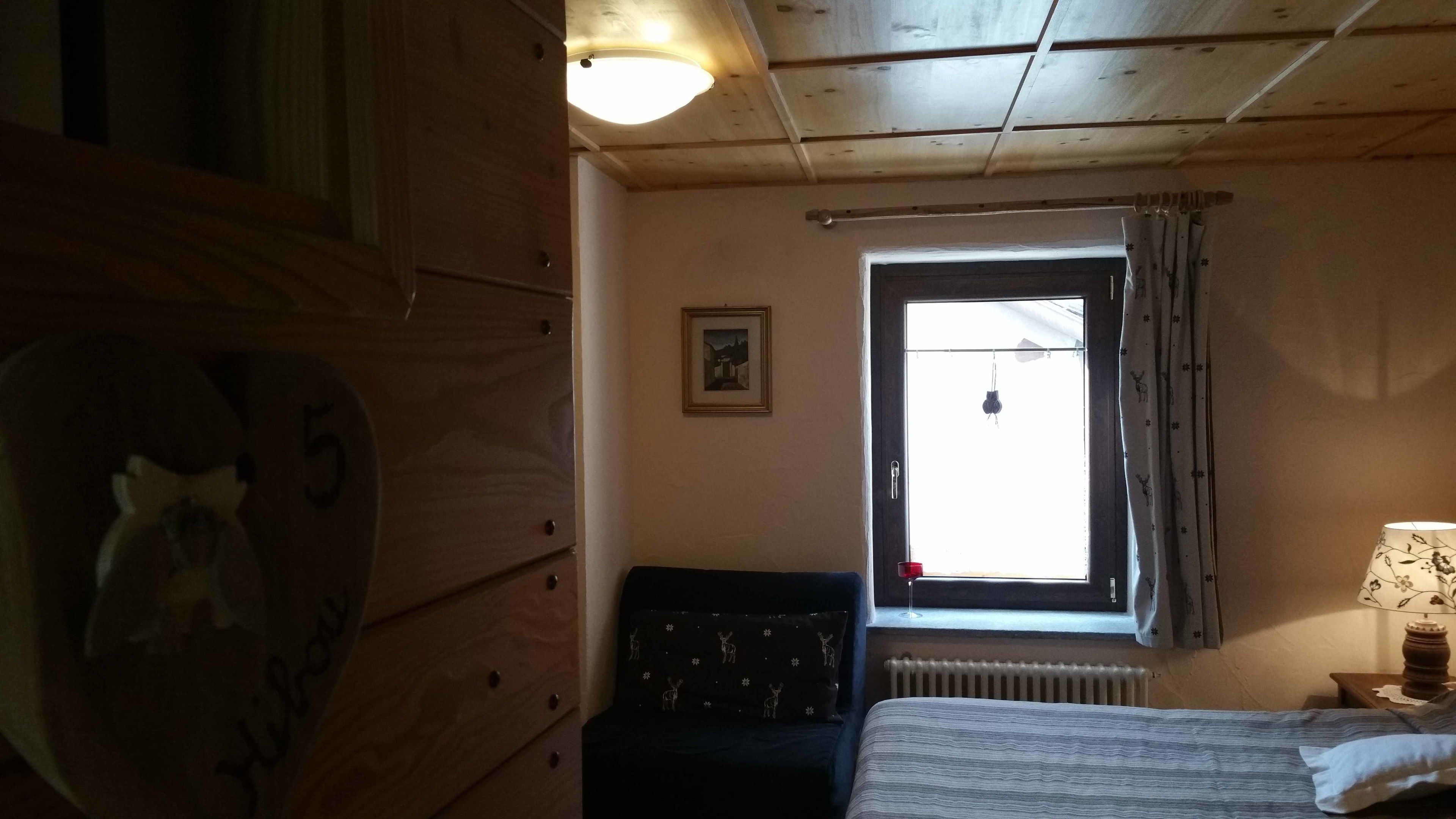 A wooden room with a bed, a chair, a radiator, a window with curtains, and a framed picture on the wall.