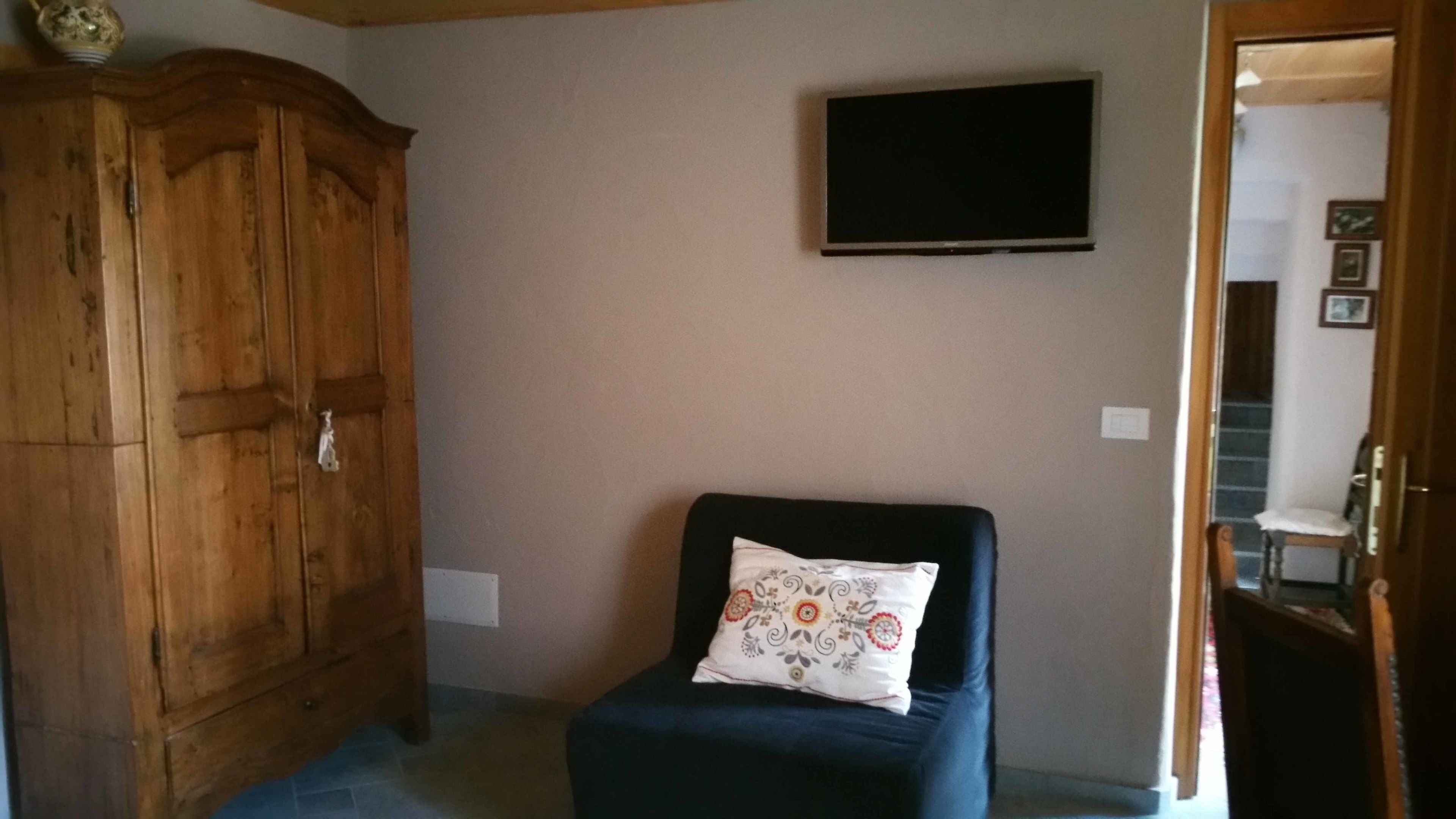 Ein Zimmer mit einem Holzschrank, einem schwarzen Sessel mit gemustertem Kissen und einem an der Wand befestigten Flachbildfernseher.