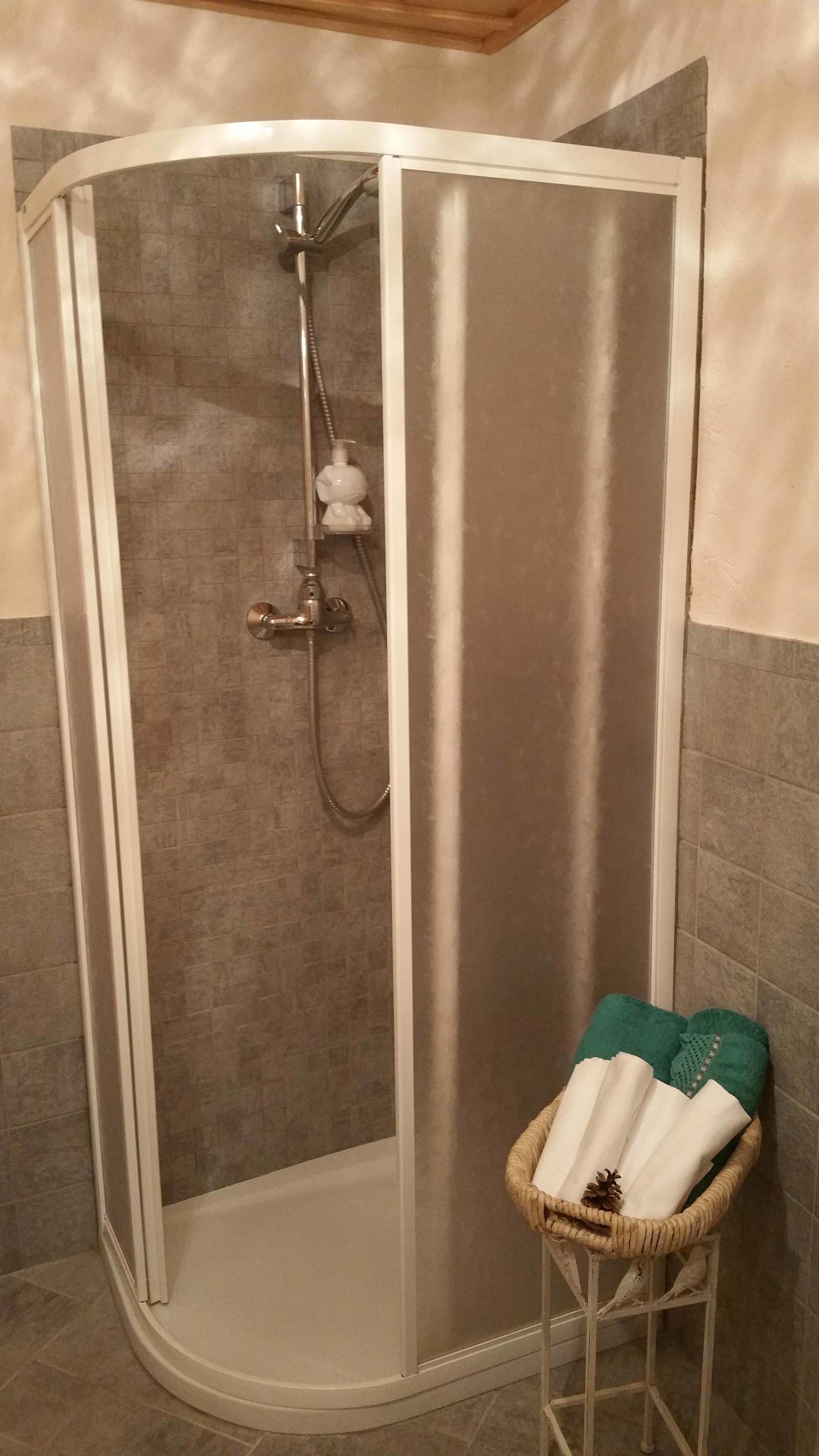 Doccia d'angolo con porta in vetro, soffione a mano e cesto portasciugamani con asciugamani.