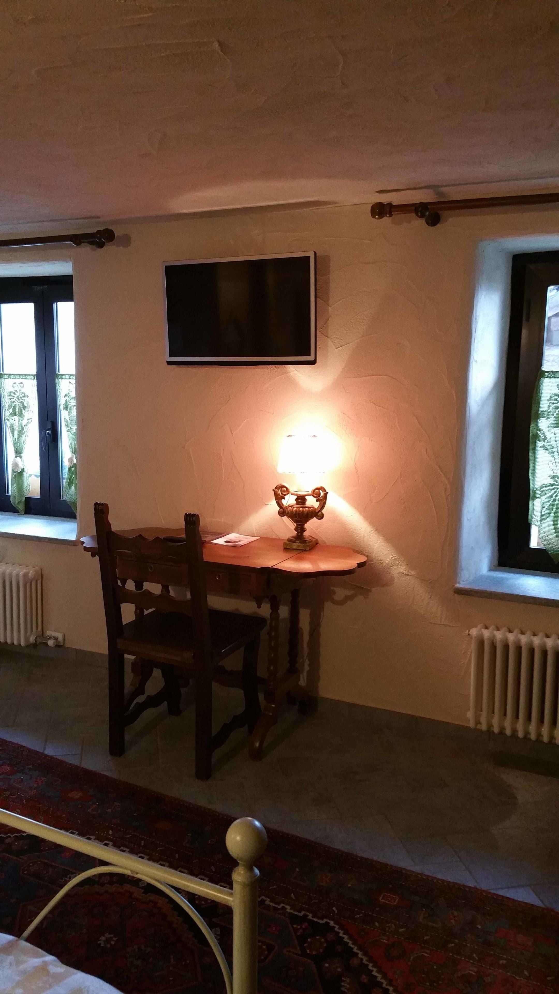 Una habitación con una silla de madera, una mesa con una lámpara, un televisor montado en la pared y una ventana con cortinas.