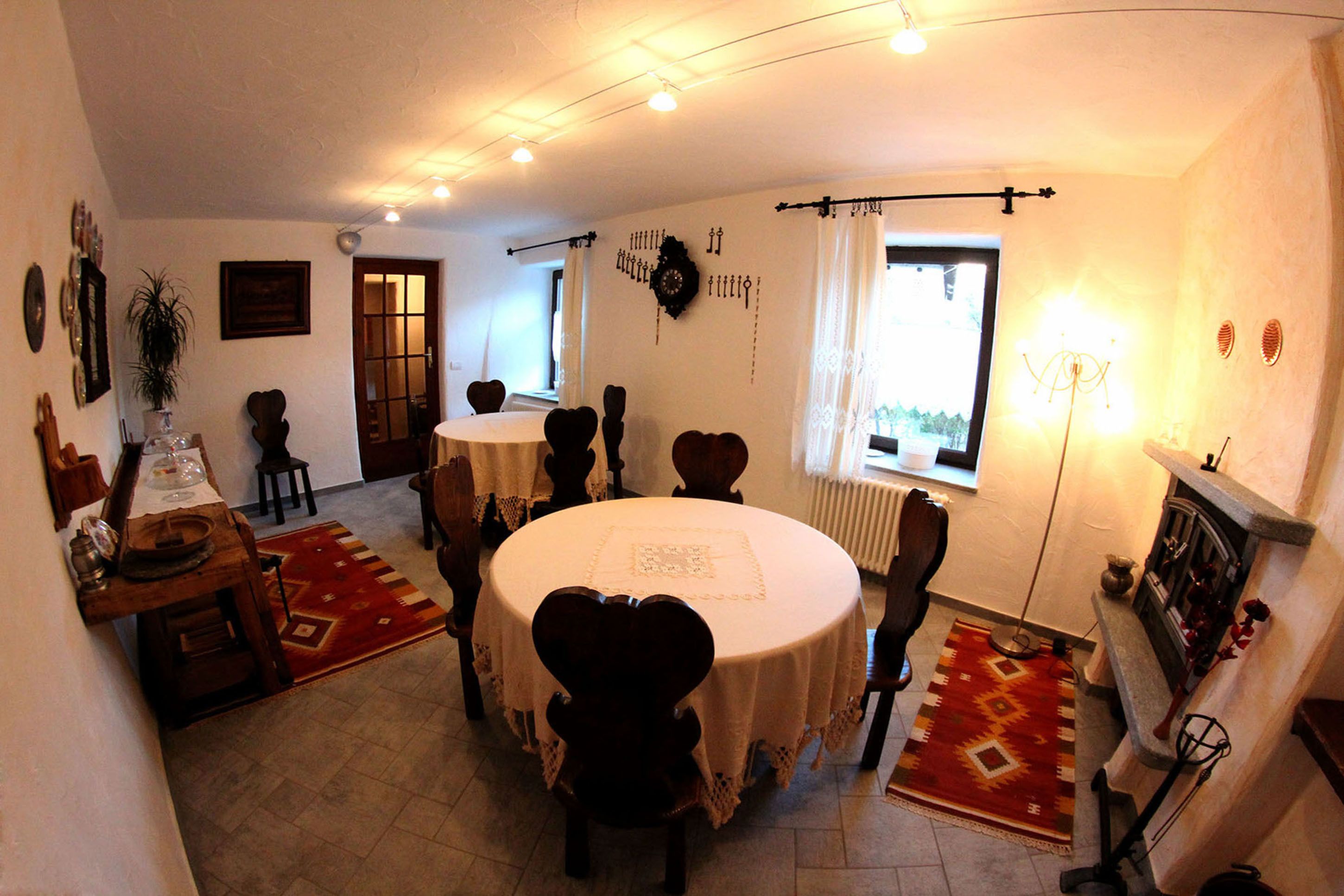 Un comedor con una mesa redonda, sillas y una alfombra roja. Se ve una ventana con cortinas, un reloj de pared y una chimenea.