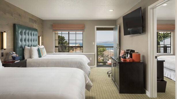 A hotel room with two beds, a TV, a balcony with a view, and a desk with a coffee machine. - Wave Street Inn (Monterey)