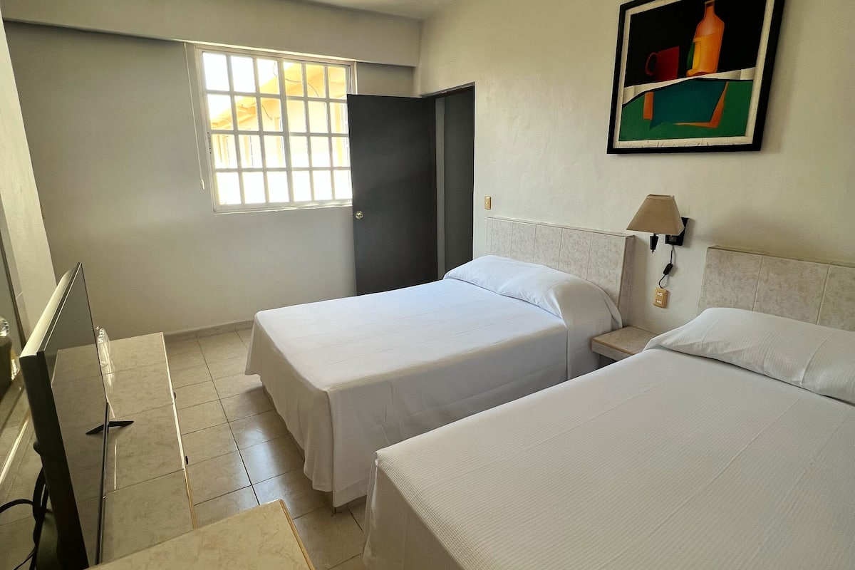 A hotel room with two beds, a window, a door, a painting, and a lamp.