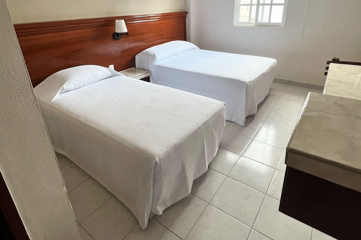 A hotel room with two beds, a nightstand, a window with blinds, and an air conditioning unit.