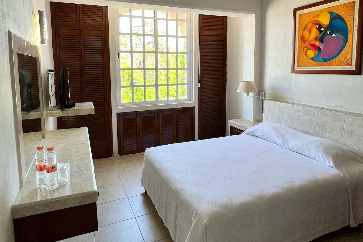 A hotel room with a bed, a desk with a mirror, a window with shutters, and a painting on the wall.