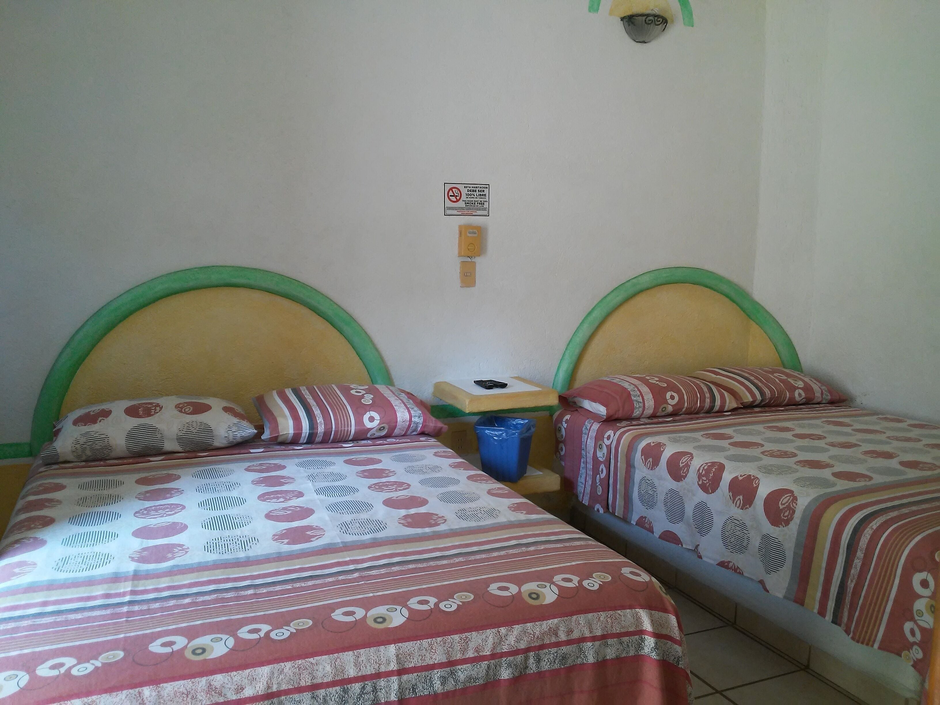 A room with two beds, each with a green headboard and patterned bedding. There is a small table with a blue trash bin and a wall-mounted light fixture.