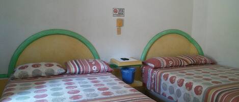 Habitación con dos camas, cada una con cabecero verde y ropa de cama estampada. Hay una mesita con un cubo de basura azul y una lámpara fijada a la pared.