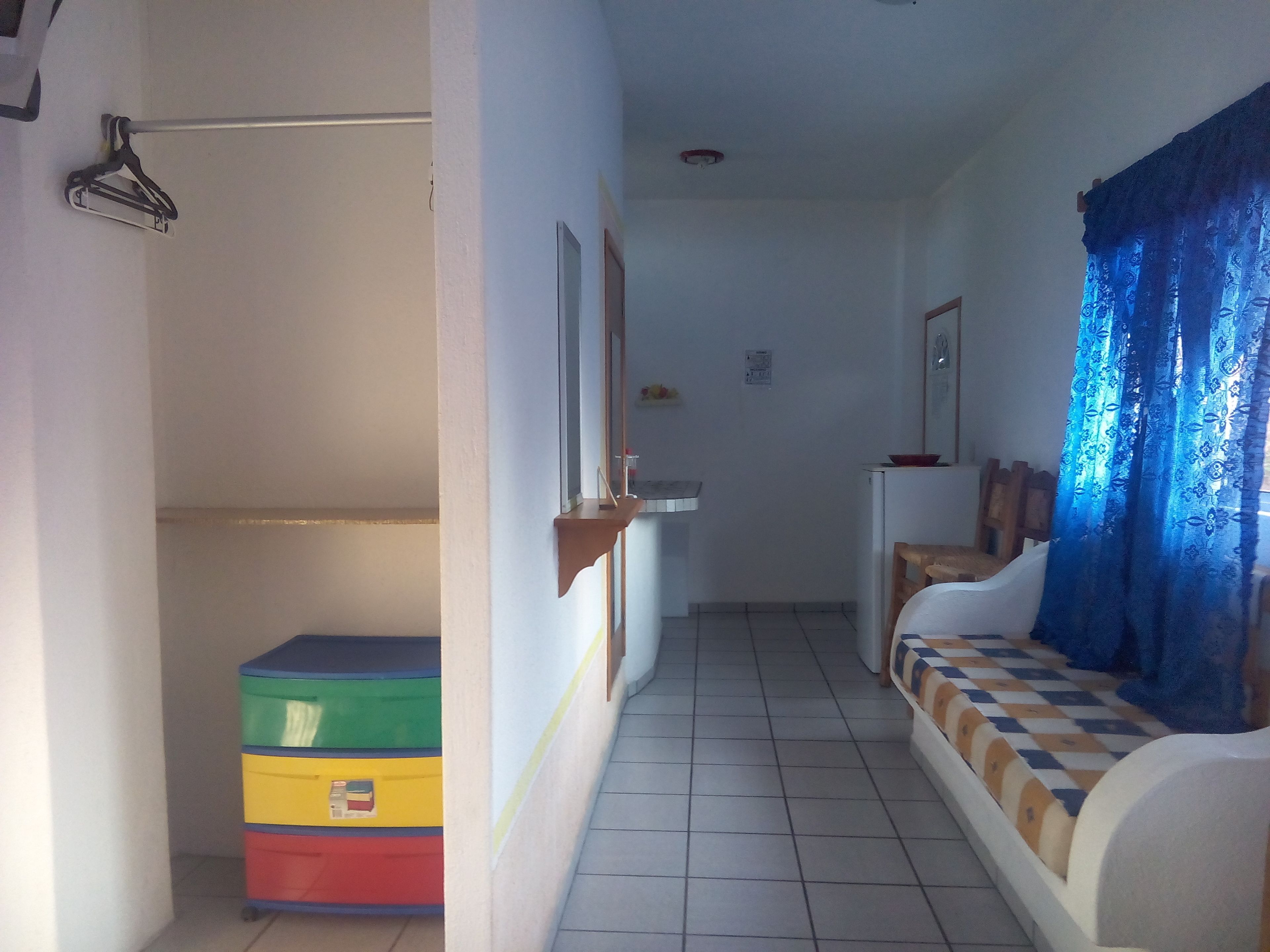 Un pasillo estrecho con suelo de baldosas, un mueble de almacenamiento colorido, un banco blanco con cojín estampado y una cortina azul.