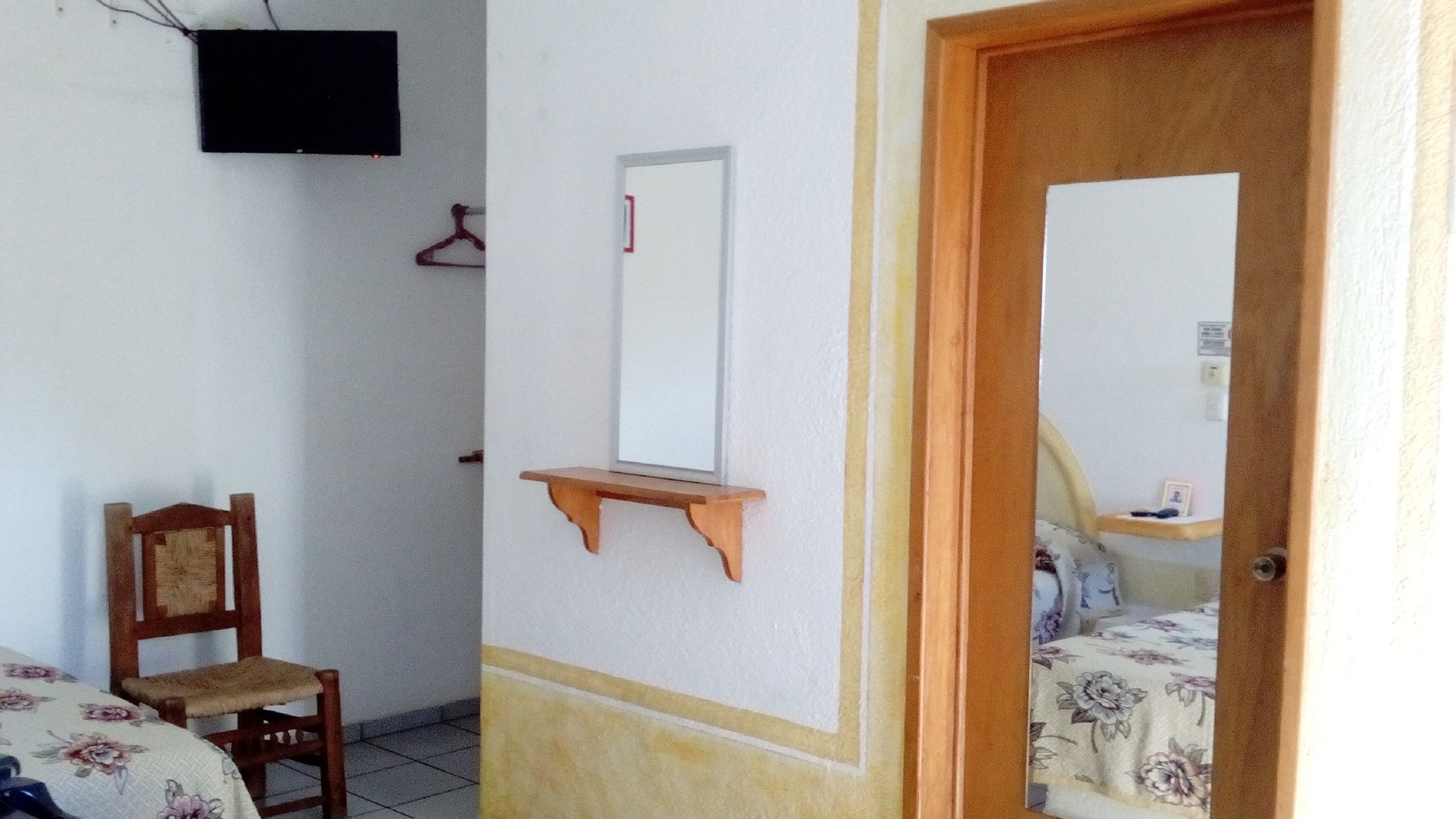 A room with a bed, a wooden chair, a small wooden shelf, a television mounted on the wall, and a door leading to another room.