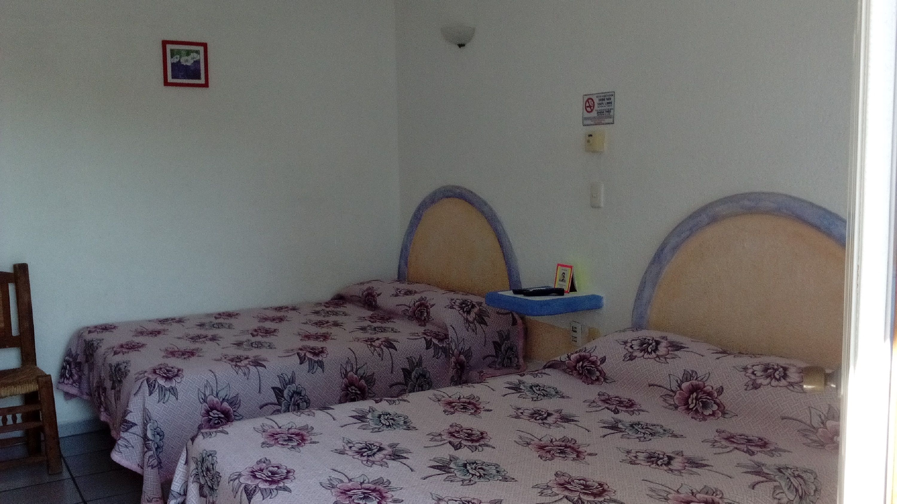 A room with two single beds with floral bedding, a small bedside table, and a wall-mounted shelf.