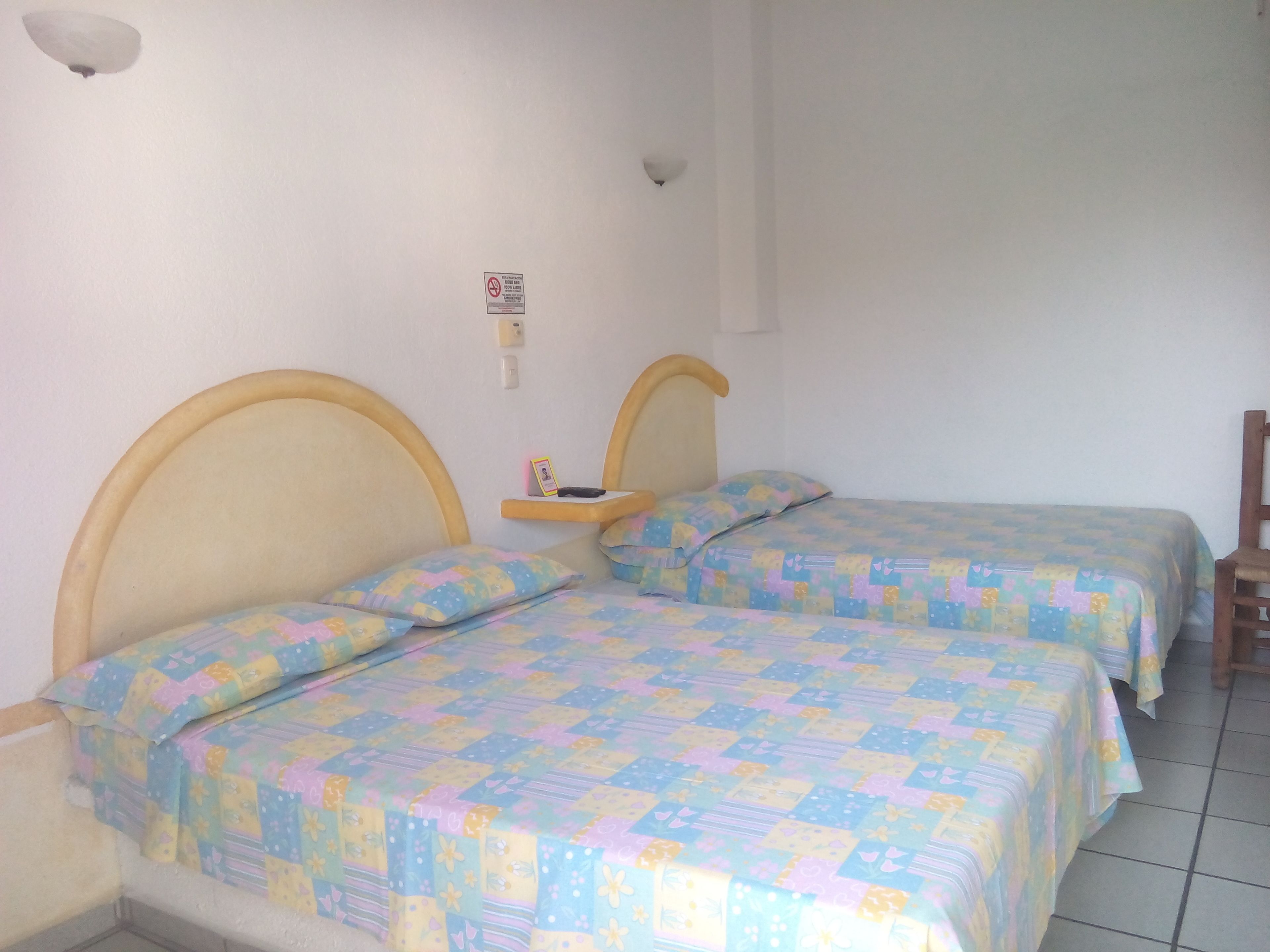 Una habitación con dos camas, cada una con cabecero de madera y colcha estampada. Hay un estante pequeño con una foto y un control remoto en la pared, entre las camas.