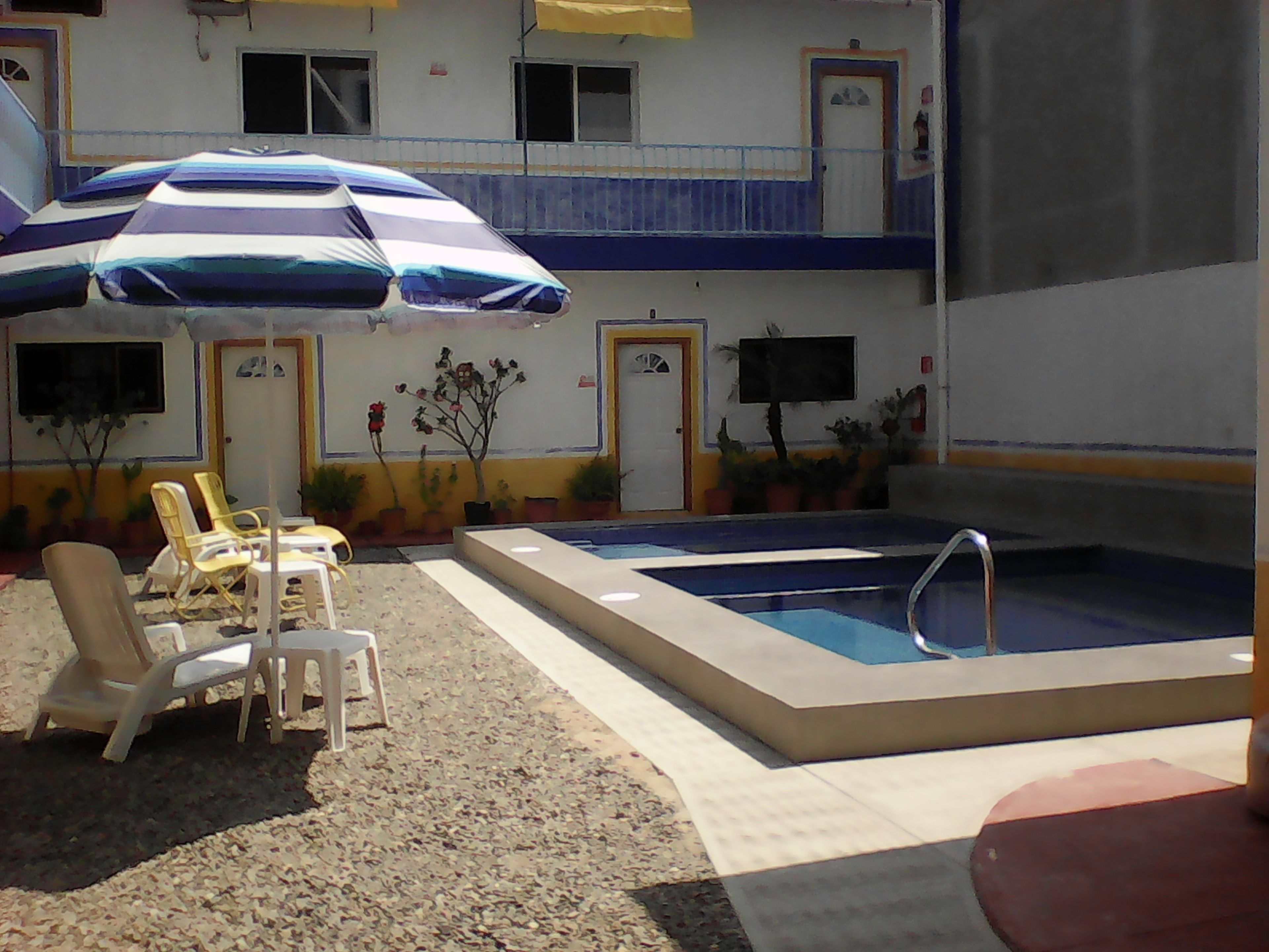An outdoor pool area with lounge chairs and an umbrella, surrounded by potted plants and a building with multiple doors and windows.