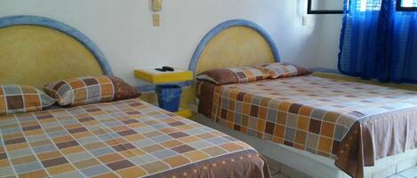 A hotel room with two beds, each with a headboard and a patterned bedspread. There is a small table with a trash bin and a sign on the wall.