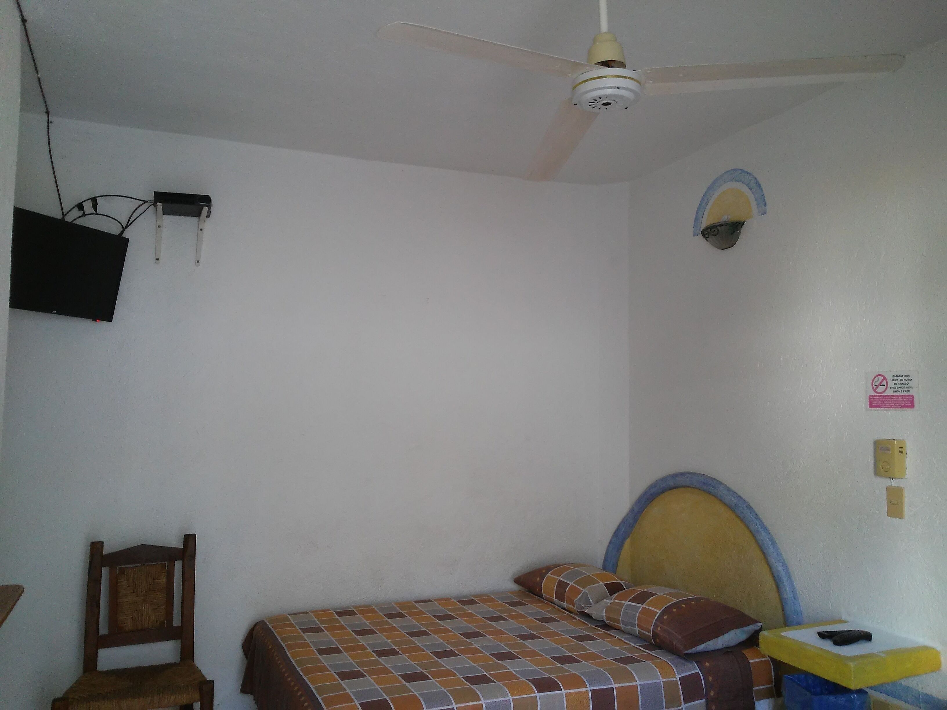 Un dormitorio con cama, ventilador de techo, televisor montado en la pared y una mesita de noche con lámpara.