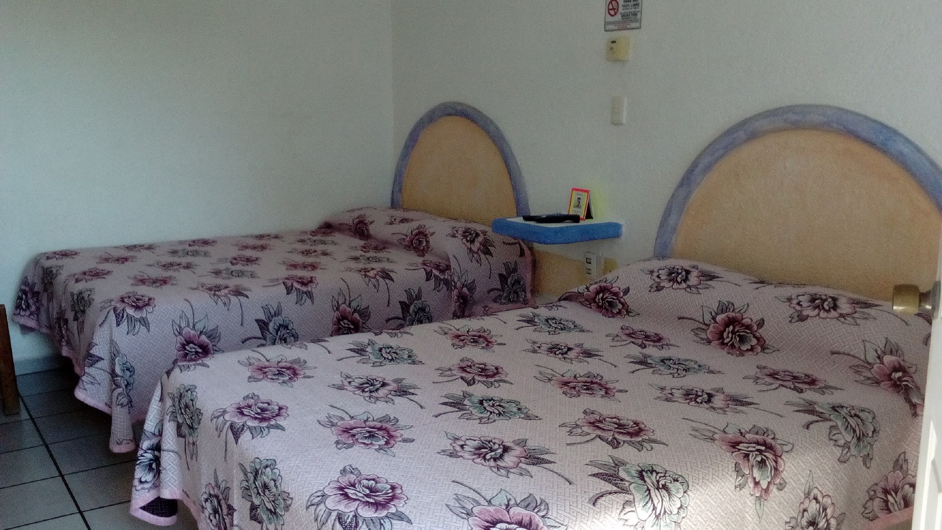 A room with two beds, each with floral-patterned bedding and wooden headboards. There is a small blue shelf between the beds.