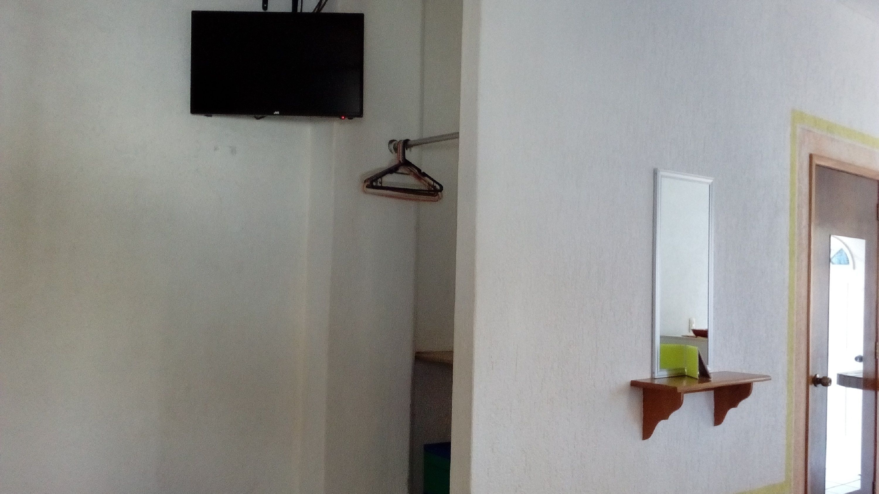 A room with a flat-screen TV mounted on the wall, a wooden door frame, and a small shelf with a mirror and a green object.
