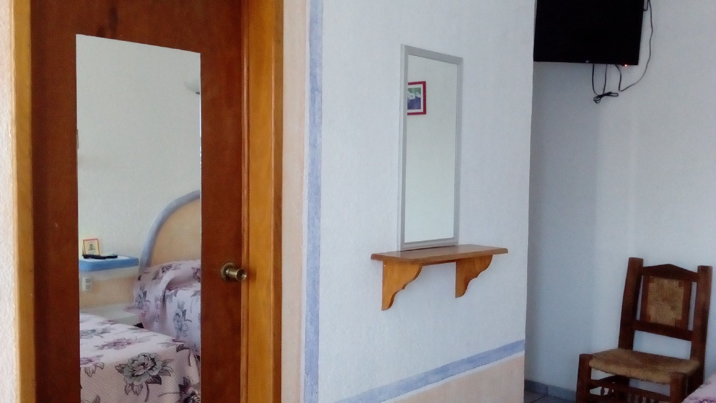A bedroom with a wooden door, a bed with floral bedding, a bedside table, a small shelf, a chair, and a mounted TV.