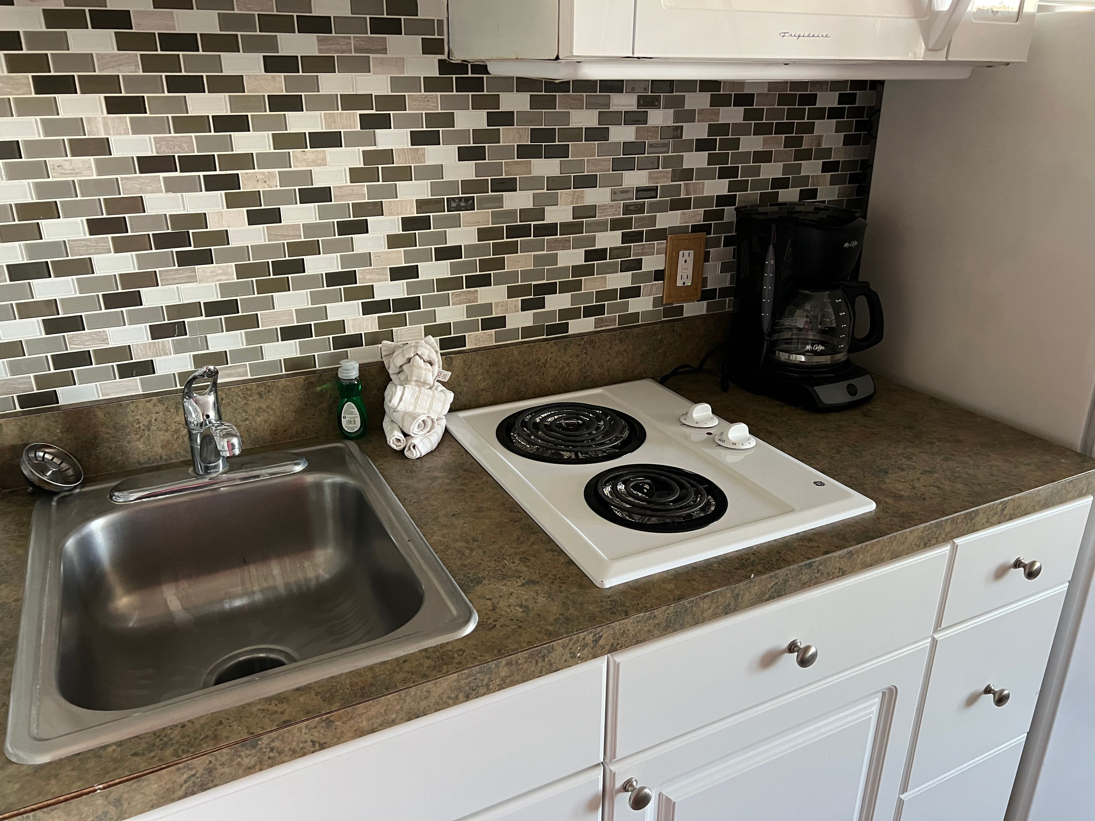A kitchen with a stainless steel sink, a white stove, a coffee maker, and a tiled backsplash.