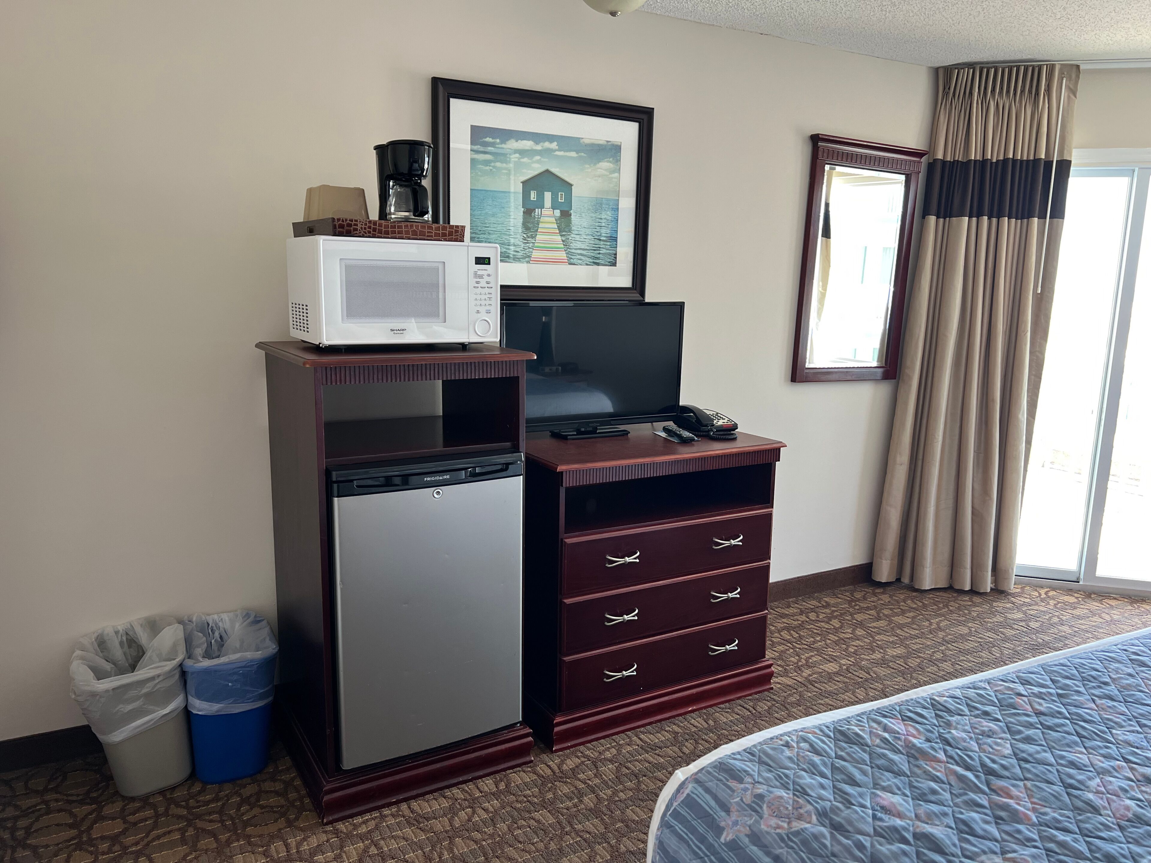 Ein kleines Hotelzimmer mit Bett, Minibar, Fernseher, einem gerahmten Bild und einem Fenster mit Vorhängen.