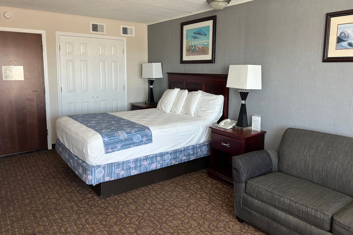 Une chambre d’hôtel avec un lit, deux tables de chevet avec des lampes, un fauteuil gris et une porte.