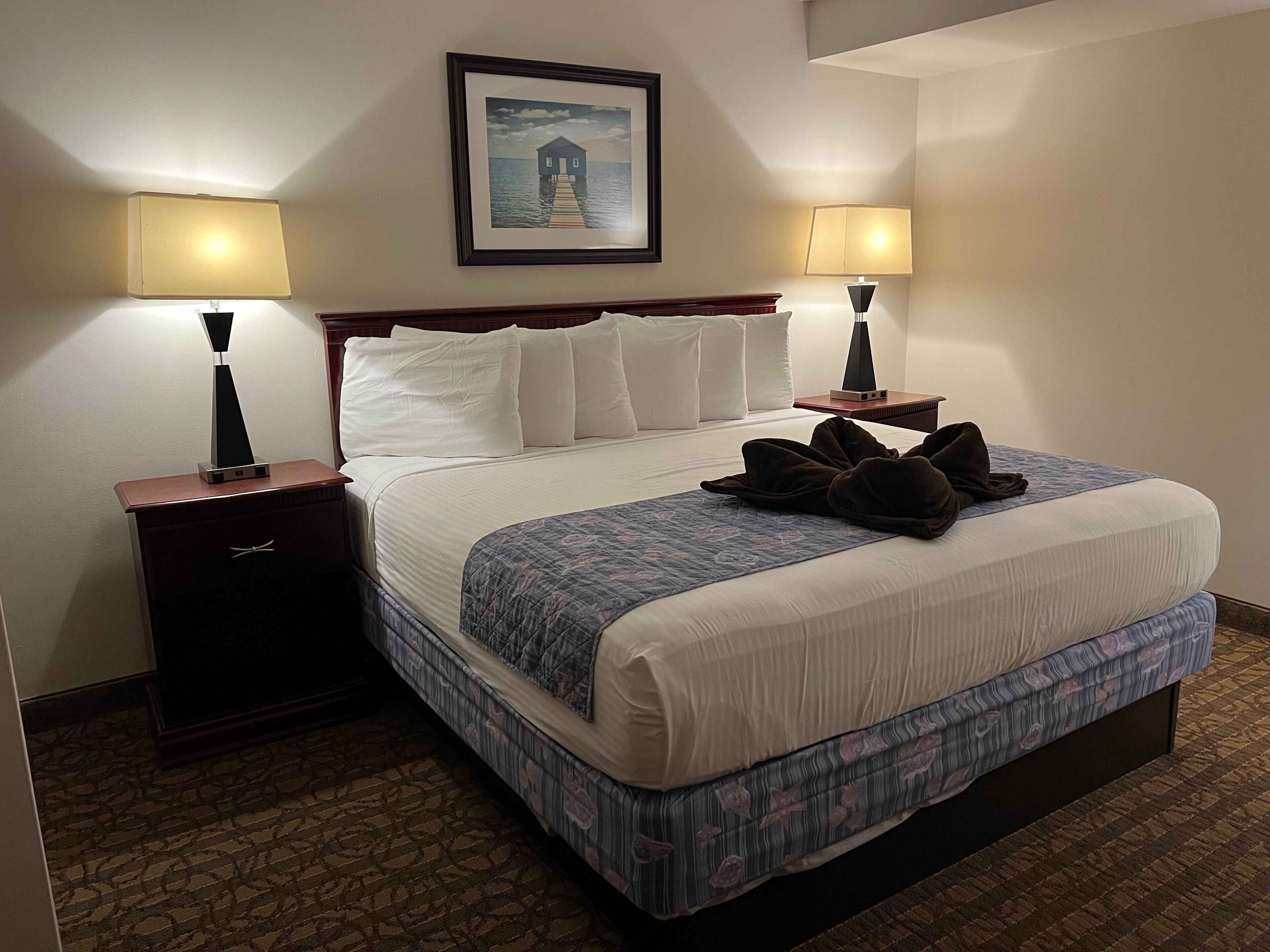 A neatly made bed with a blue and white patterned bedspread, two bedside lamps, a framed picture of a house on water, and a wooden nightstand with a drawer.