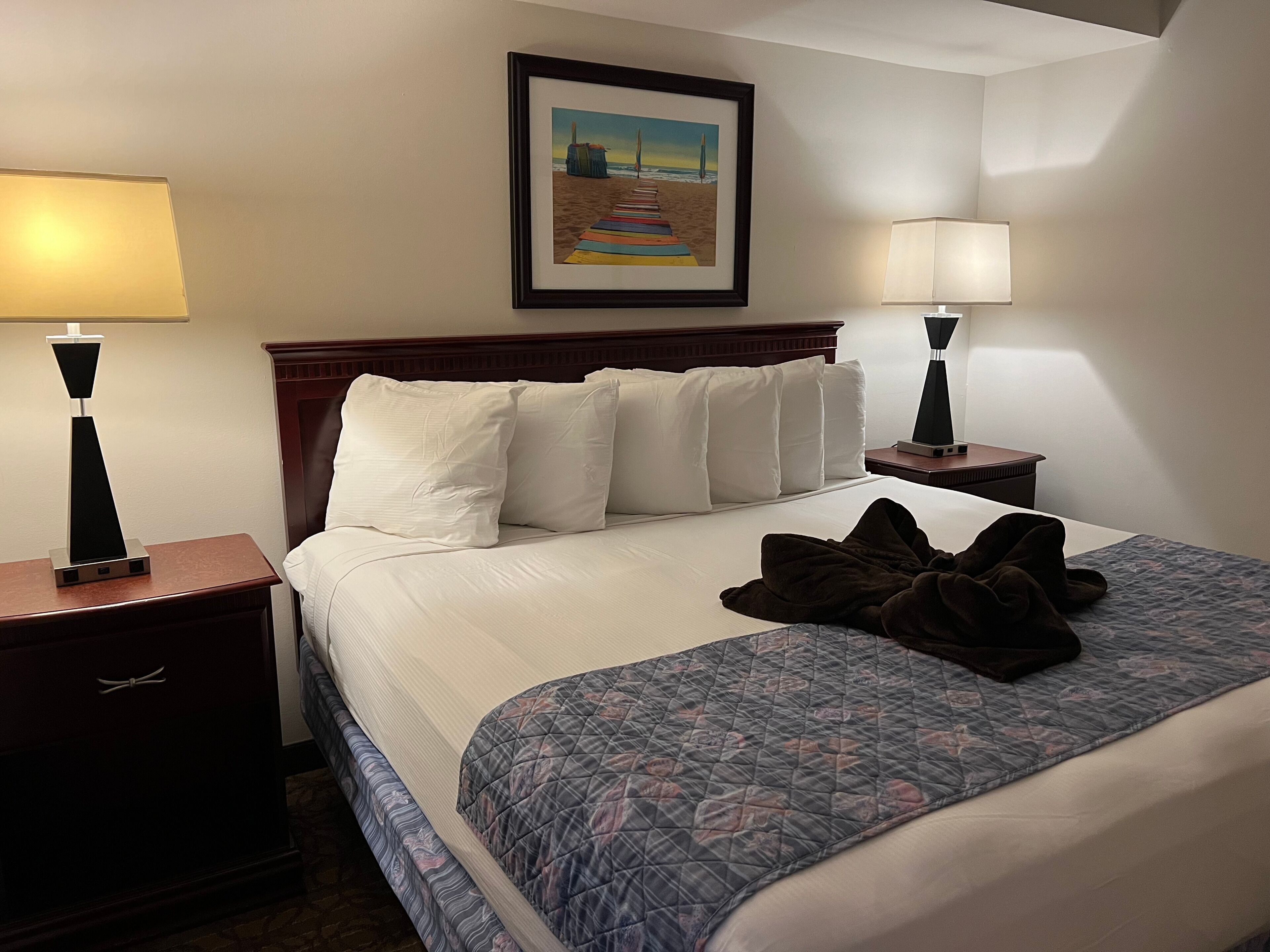A hotel room with a neatly made bed, two bedside lamps, a framed beach painting on the wall, and a wooden headboard.