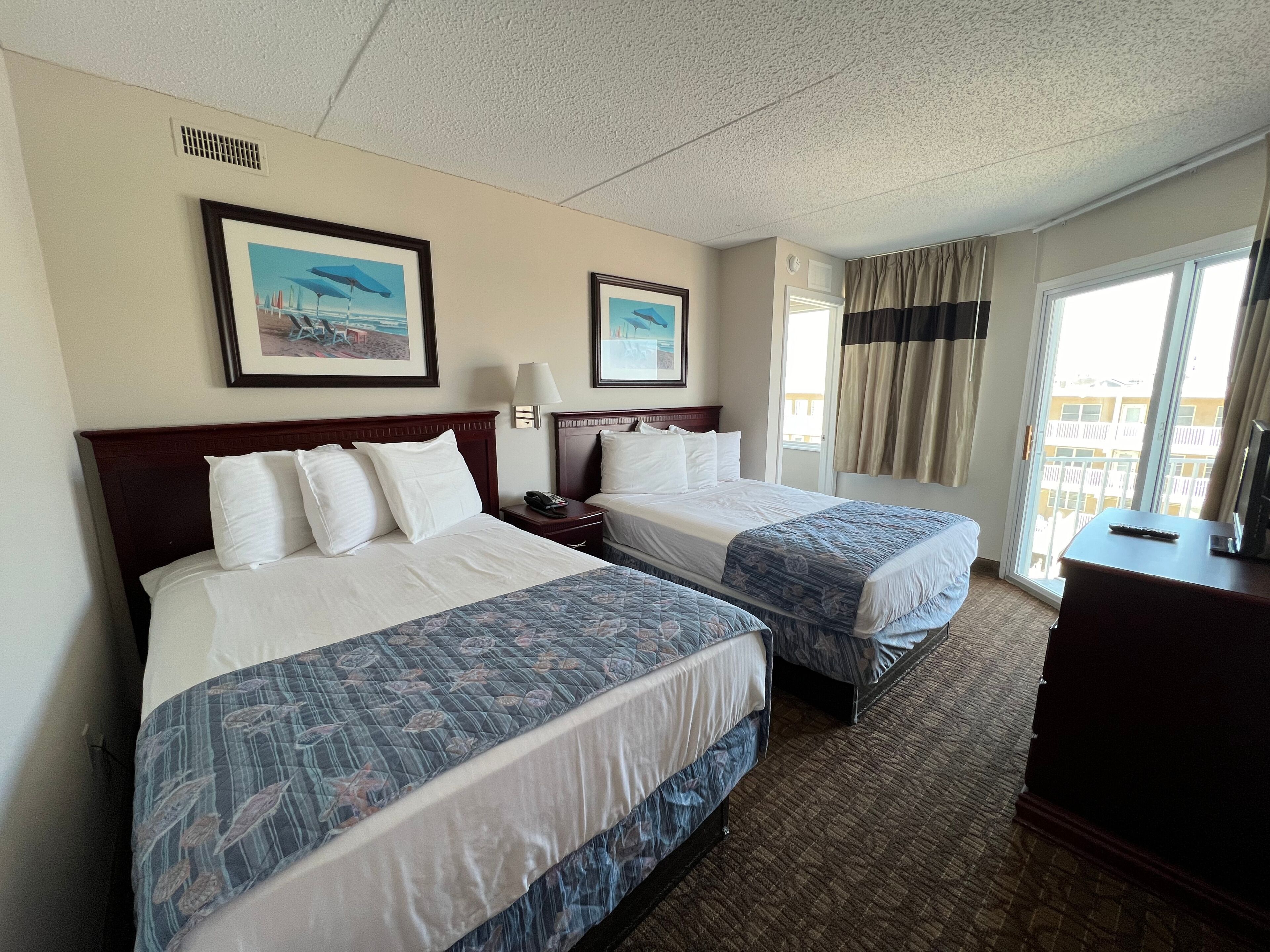 A hotel room with two beds, a nightstand with a lamp, a desk with a remote control, and a window with curtains.