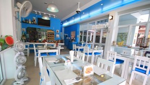 A restaurant with blue walls, white tables, and chairs. There are fans, a bar area, and a large window. - Karon Sea Side (Karon)