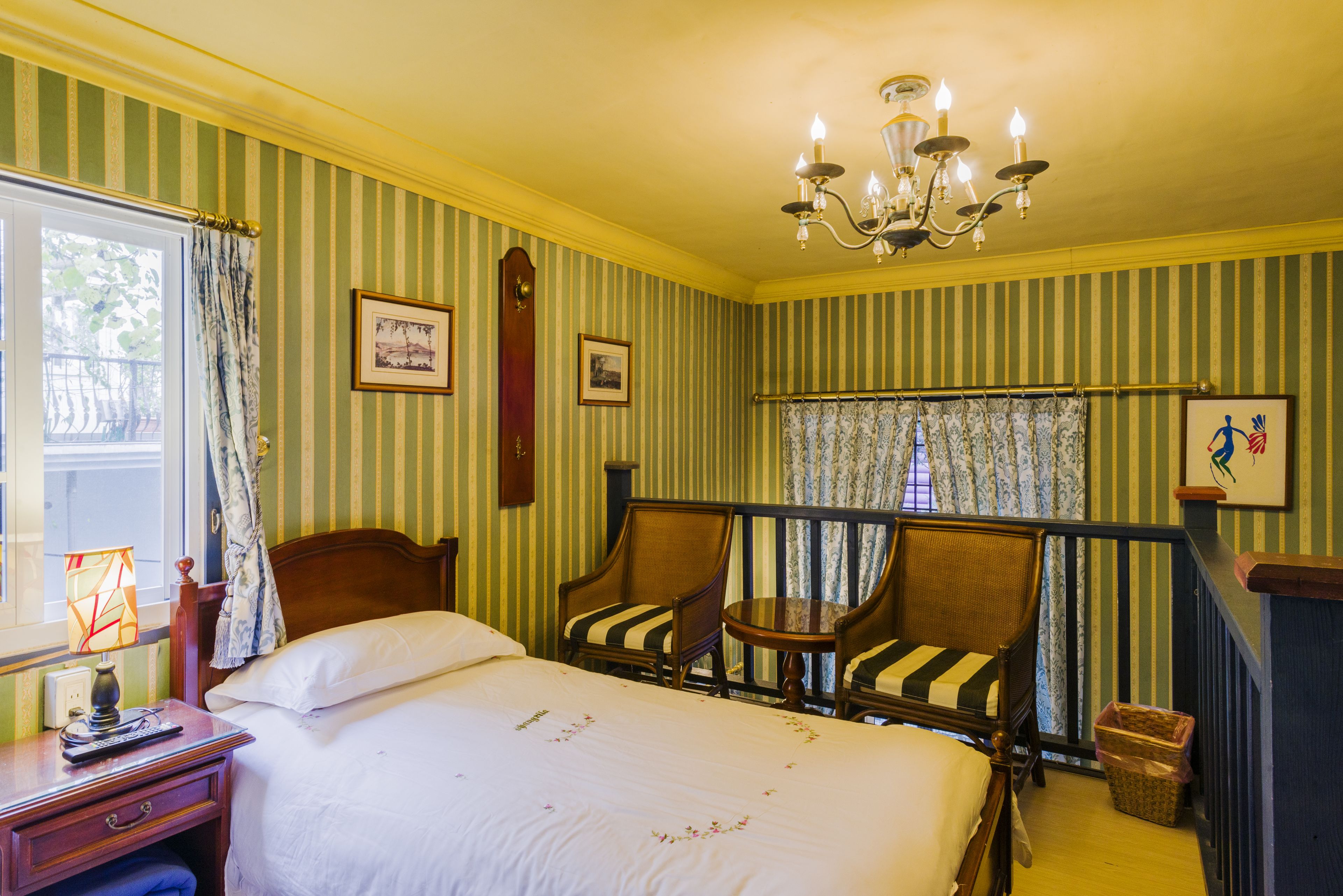 A hotel room with a bed, two striped armchairs, a small table, a chandelier, and a window with curtains.