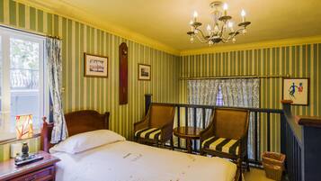 A hotel room with a bed, two striped armchairs, a small table, a chandelier, and a window with curtains.
