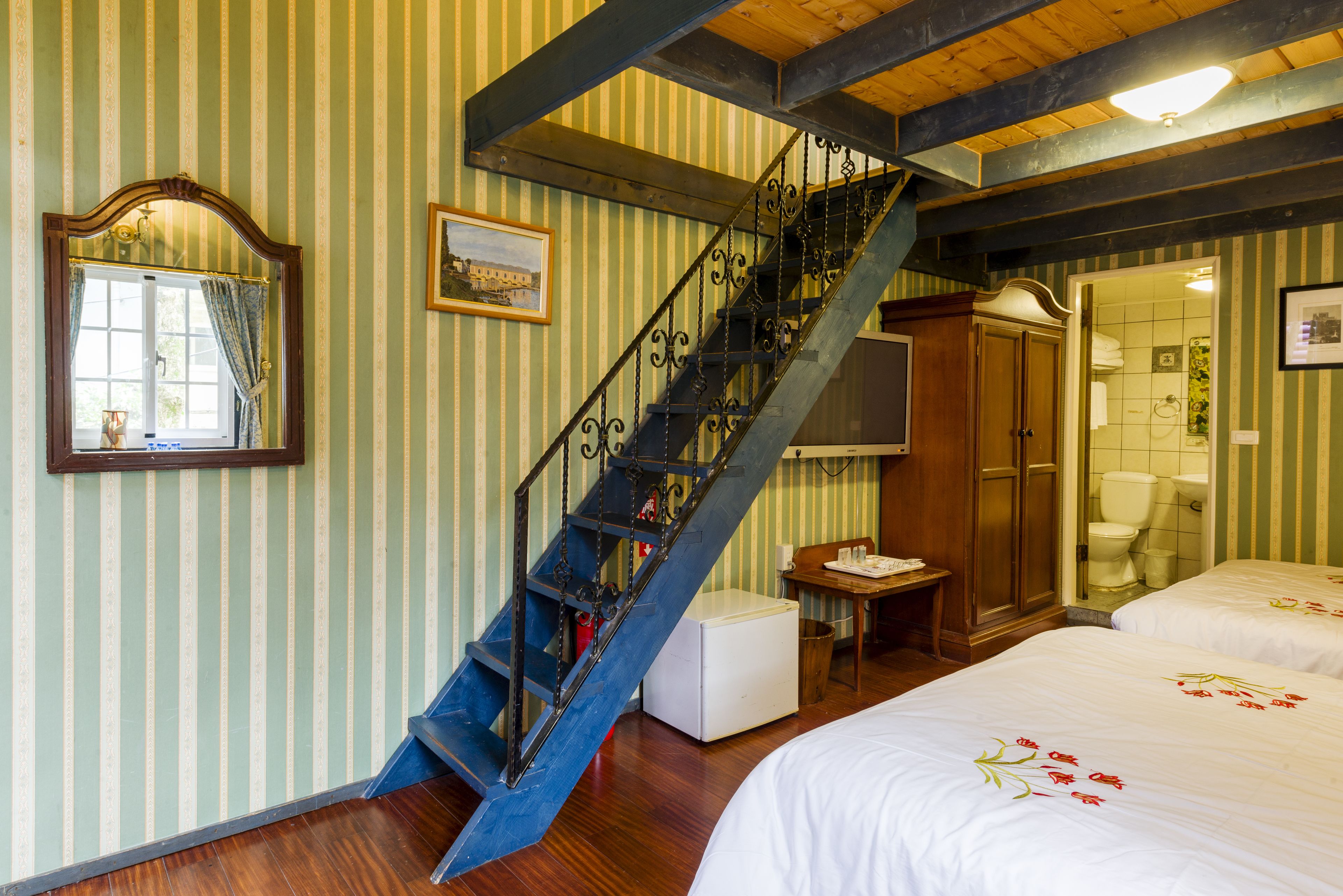 A room with a wooden staircase, a bed with floral bedding, a wooden desk, and a mirror with a picture frame.