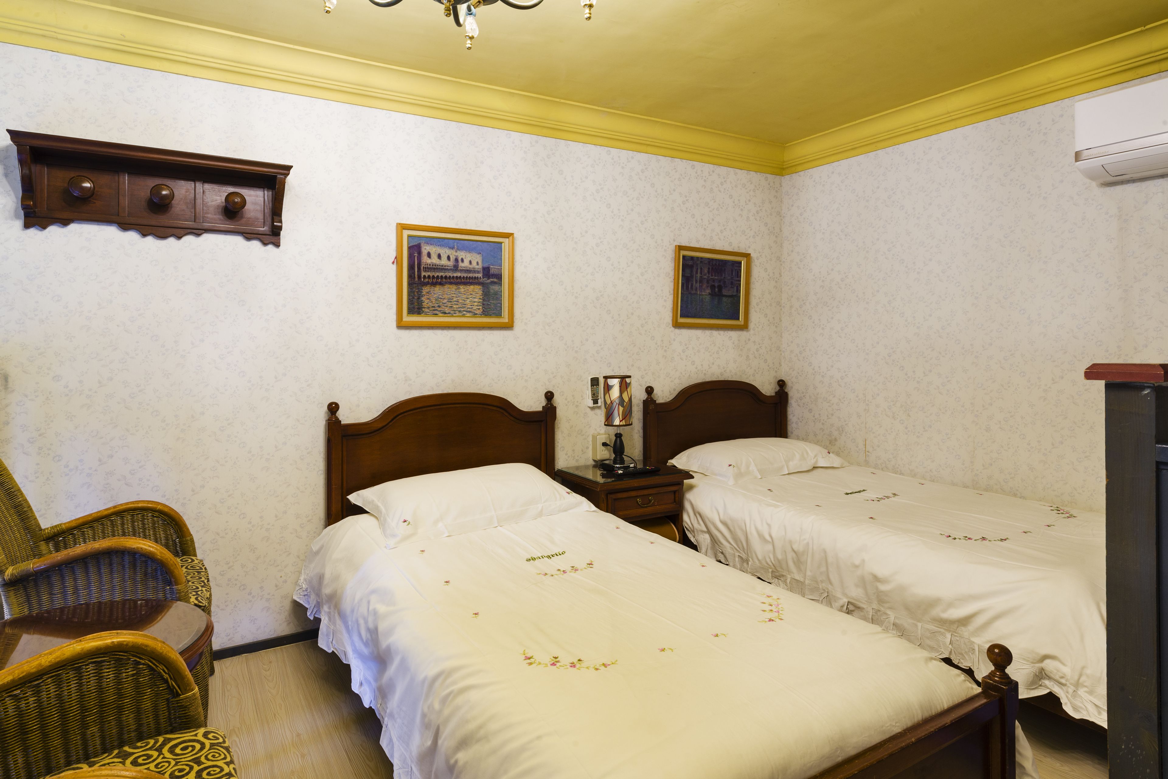 Una habitación de hotel con dos camas individuales, una mesita de noche de madera, un sillón de mimbre y un estante de madera.